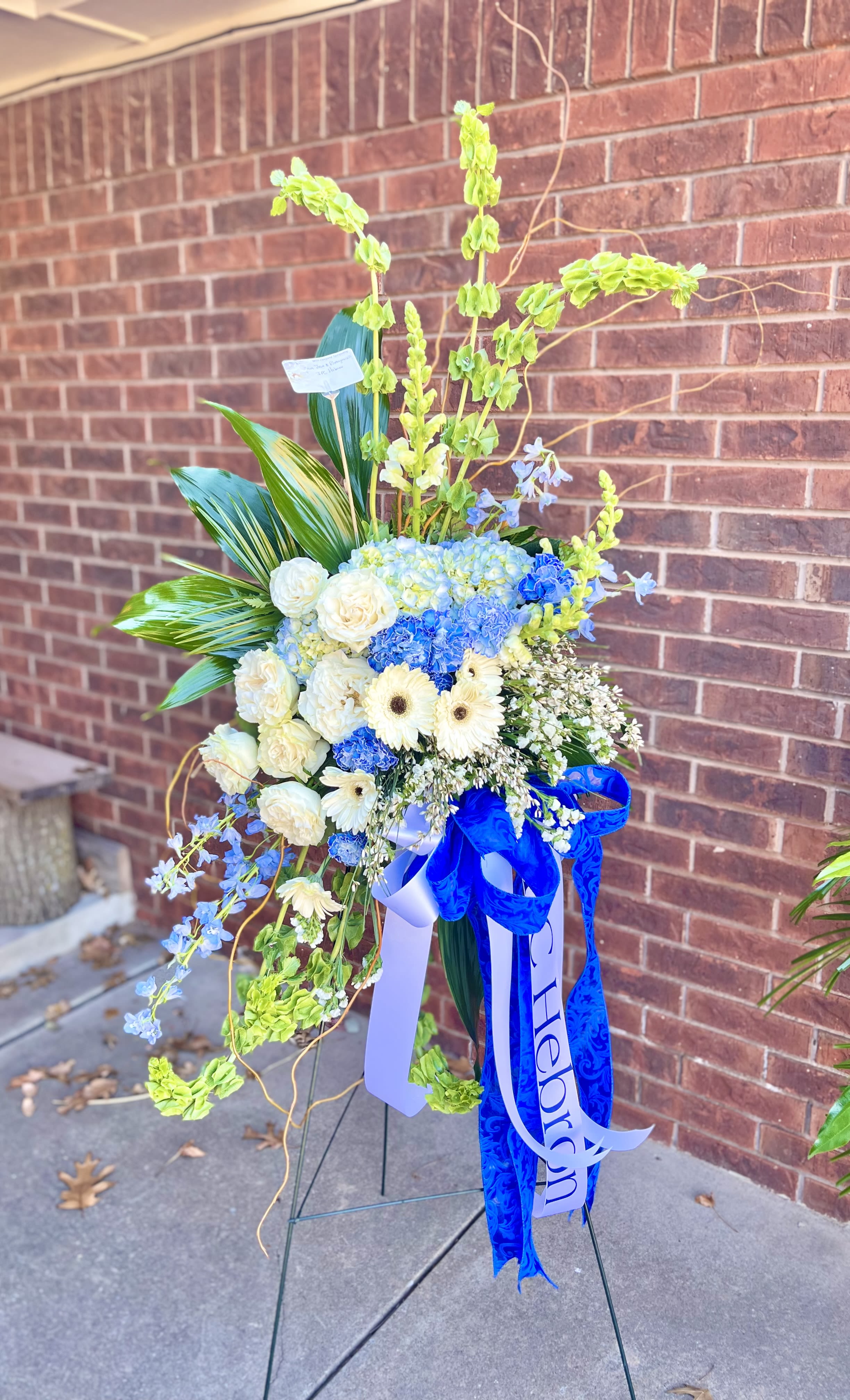 Falling Waters - A tall, graceful standing spray that reads as a serene tribute, Falling Waters is a sympathy flower arrangement designed to convey quiet support at a memorial or funeral. The vertical composition and cascading lines give the piece a feeling of gentle movement-perfect for honoring a loved one or offering comfort to someone grieving.  This arrangement centers on a dense cluster of blue and white blooms-roses, hydrangeas, gerbera daisies and complementary seasonal flowers-set against airy green and pale blue flowering stems that arch upward and outward. Rounded blossoms mingle with long, elegant lines to create a layered, formal silhouette. A large blue ribbon and white ribbon drape from the front and a printed memorial ribbon is included; the spray is displayed on a sturdy metal easel for ceremony-ready presentation.  Made to comfort the person you're thinking of, Falling Waters sends a message of peace and remembrance at a funeral or memorial service. The standing spray arrives assembled and ready to place on-site; to preserve presentation, keep out of direct sun and away from heating vents until the service.