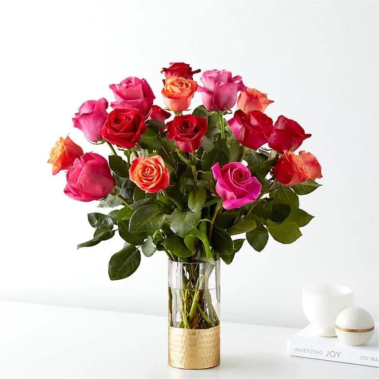 Vivid Rose Story - A lush cluster of roses in vivid pinks, reds, peaches and pops of orange and white creates an instantly celebratory feel-perfect as a birthday bouquet, anniversary roses, or a romantic Valentine's Day surprise. The Vivid Rose Story reads lively and heartfelt; the person you're thinking of will feel brightened and truly noticed by this saturated, classic rose composition.  Tightly gathered and layered at varying heights, the round silhouette sits upright in a tall clear glass vase with a textured gold band at the base that lends a polished, contemporary finish. The mix of warm hues and leafy green accents gives visual depth and a joyful contrast while keeping the arrangement elegant and restrained.  Ideal for sending as a thank-you, a love note, or to mark a special milestone, this arrangement arrives ready to display-no extra styling required. Standard vase dimensions and care: trim stems and refresh water every 2-3 days to keep the roses looking their best through the week.