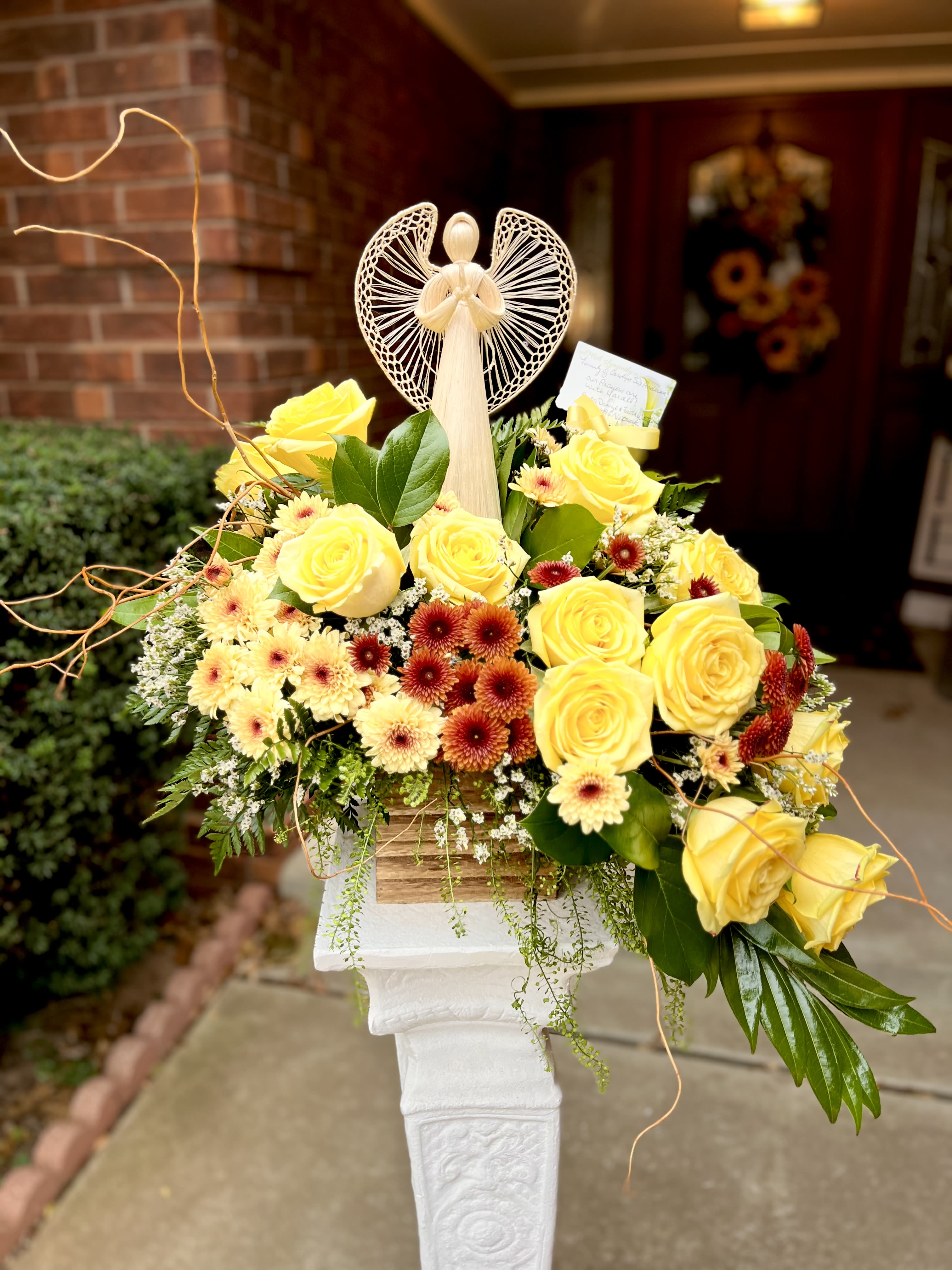 Golden Embrace - Sunny yellow roses clustered with cream daisy-like chrysanthemums and deep red accents create a warm, uplifting sympathy arrangement or thank-you gift that feels like a gentle hug. As a flower arrangement for memorials, get-well wishes, or to celebrate someone who brings light into your life, this piece centers on comforting color and thoughtful detail to make the recipient feel truly seen.  The design is full and layered: bright yellow roses mingle with small cream and deep red blooms, set against fresh greenery and spilling naturally from a rustic wooden container. A delicate angel figurine rises from the center, adding a quietly reverent focal point, and a keepsake card is included to carry your message. The texture of the roses and daisy-like chrysanthemums paired with trailing accents gives a hand-tended, homey look that reads as both sunny and serene.  Perfect for sending sympathy, expressing gratitude, or marking a gentle celebration, this arrangement arrives ready to display in its wooden vessel-no extra vase required. Include your personal note on the enclosed card; with basic water care (refresh water and trim stems every few days) the blooms will maintain their bright, comforting presence.