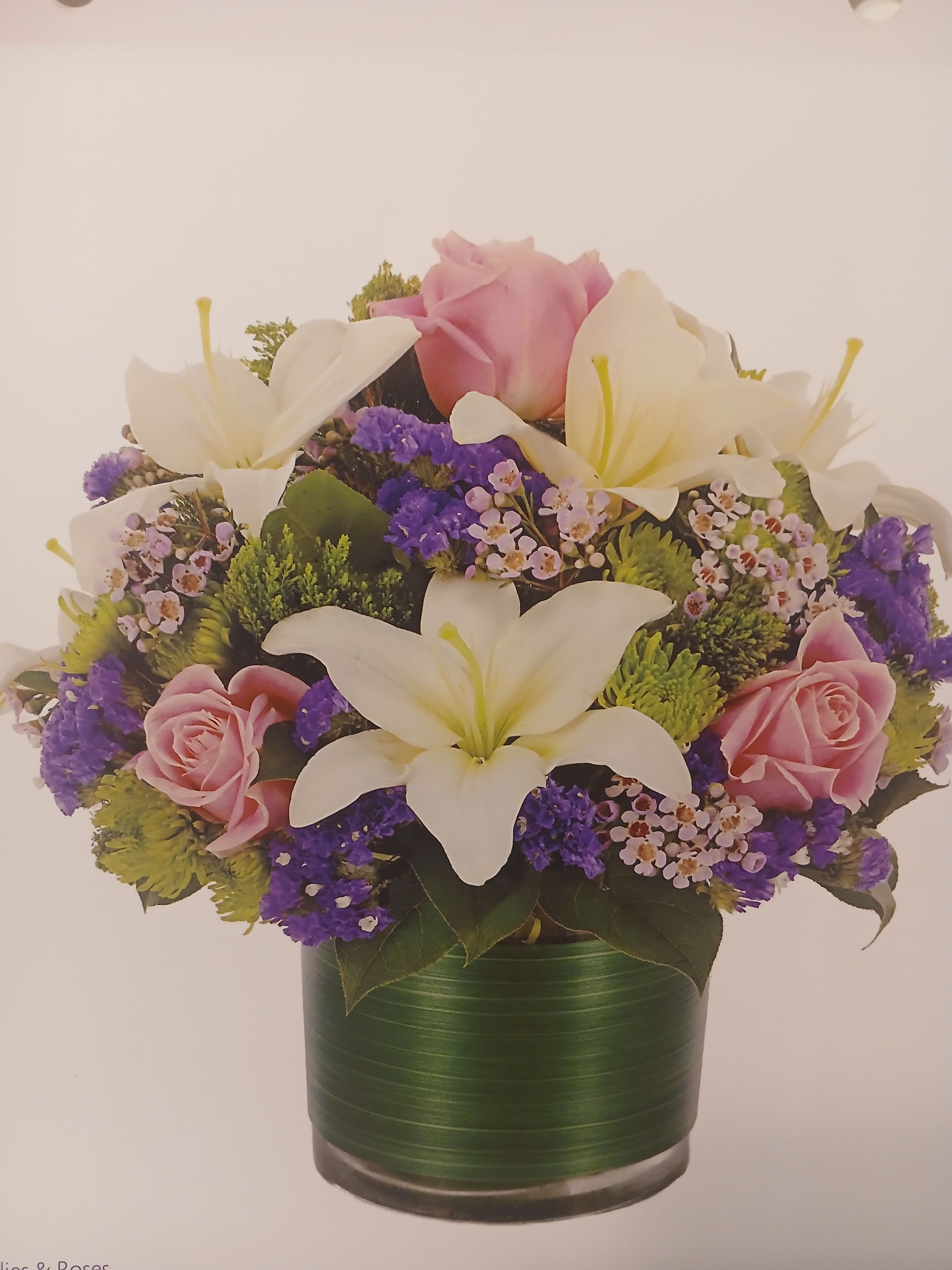 "Glorius Blooms" - Large white lilies open like soft stars at the heart of this pink, purple, white, and green arrangement, lending a fresh, elegant focal point that feels both romantic and quietly uplifting. Perfect as an anniversary bouquet, birthday surprise, or a thoughtful Mother's Day gift, these blooms also provide a gentle, respectful presence for sympathy and funeral services or for a "thinking of you" moment in spring and summer.  Soft pink roses and layered chrysanthemums nestle around clusters of small purple filler flowers, creating a rounded, full silhouette with dark foliage that makes the pale blooms pop. Arranged in a simple green vase, the composition reads as refined yet approachable-rich textures and subtle color contrasts give depth without fuss. The size is substantial enough to stand on a mantle or bedside table; stems arrive ready to display and benefit from fresh water and occasional recutting to extend vase life.  Whether you're celebrating a milestone, offering condolences, or brightening someone's day just because, this arrangement sends a sincere message of care and warmth. It's a fitting choice for women's day, congratulations, or seasonal spring and summer moments when you want the recipient to feel seen and comforted.