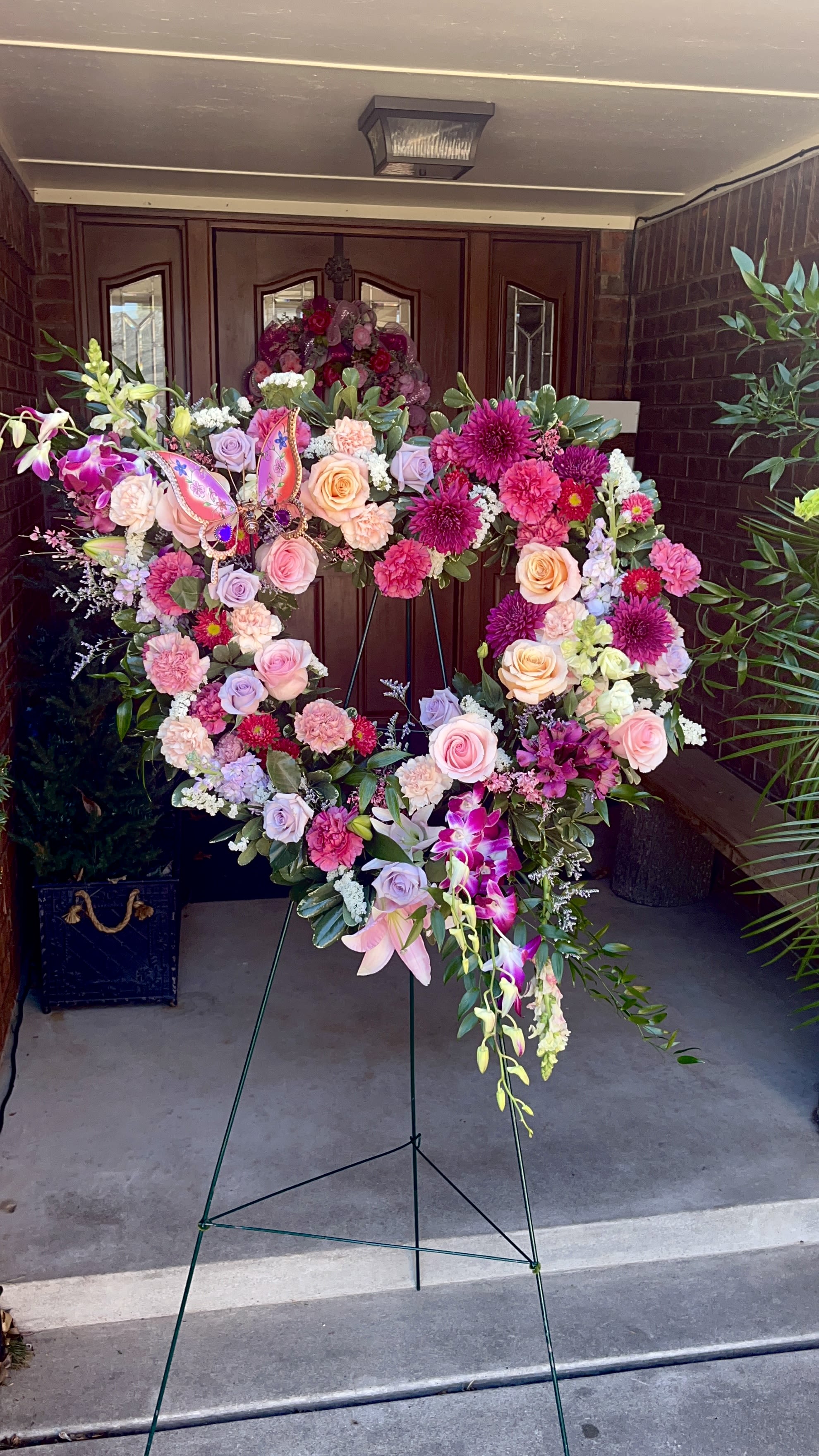 Deborah’s Heart - A tender heart of roses, lilies and orchids-Deborah's Heart is a large, standing heart-shaped sympathy arrangement designed to honor a life with grace. Perfect as a memorial tribute or for a funeral service, this floral heart conveys sympathy and remembrance through a soft palette of pink, purple, peach and white that reads both comforting and reverent for the person you're remembering.  Densely packed blooms-roses, carnations, chrysanthemums, orchids, lilies and alstroemeria-are layered to create a lush, textured surface that holds its heart silhouette on a sturdy metal easel. The elevated presentation gives the piece formal presence at a service while the decorative butterfly offers a delicate, personal accent. The combination of cool lavenders, warm peaches and classic white creates depth and a gentle visual rhythm that remains floral-forward and solemn.  Made to offer a meaningful expression of care, Deborah's Heart is suited for family tributes, memorials, and graveside services when you want someone to feel seen and remembered. Arrives ready to display on its easel; light misting of the blooms and keeping the arrangement out of direct sun will help preserve freshness during the service.