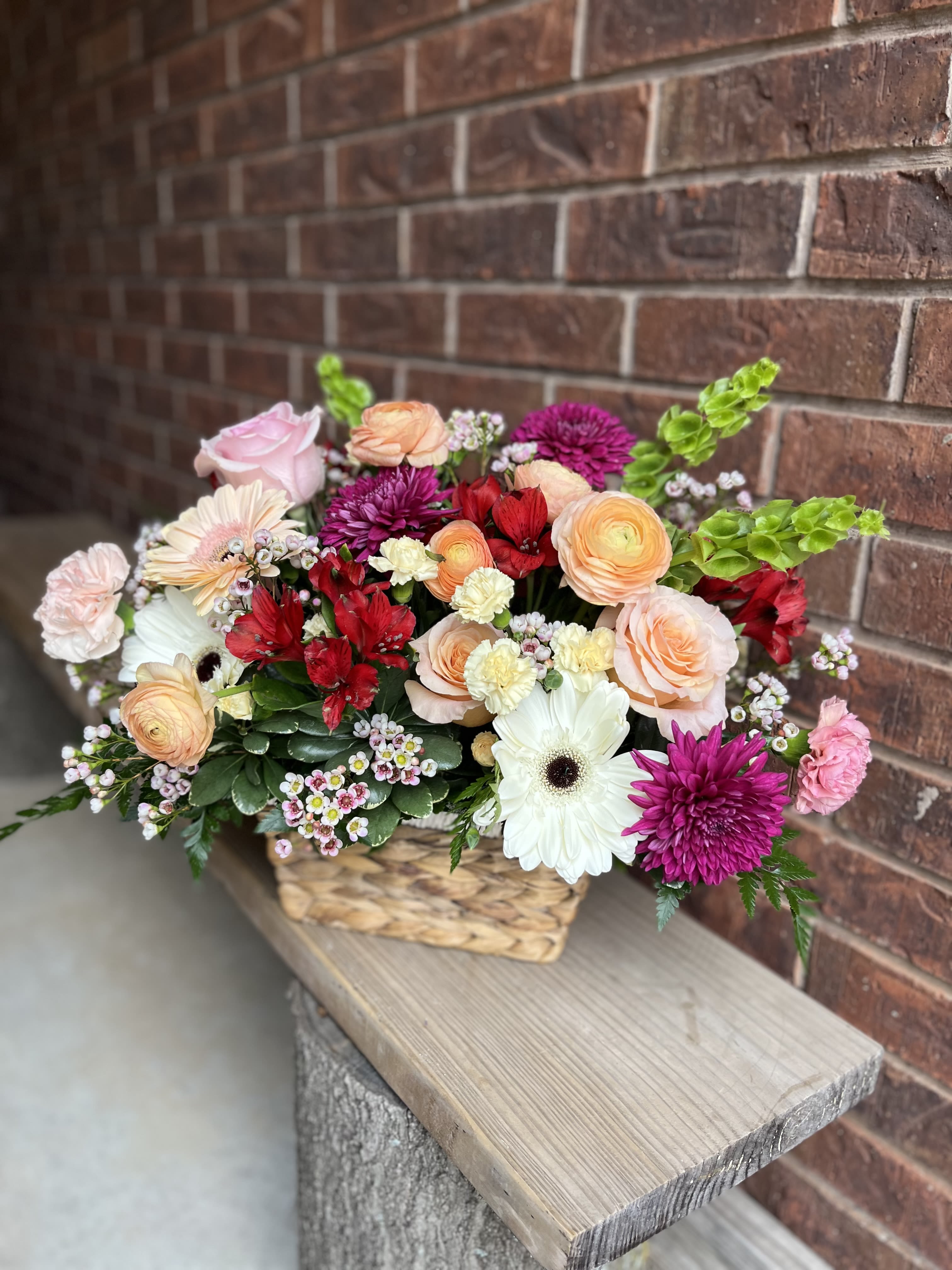 Berry Peach - A lush, hand-tied look in a low woven basket-this Berry Peach flower arrangement brings a cheerful, romantic garden feel to birthdays, anniversaries, or thoughtful just-because moments. The full, low basket design places the flowers close together so the person you're thinking of is greeted by a burst of color and texture the moment the lid is lifted.  Packed with roses, large gerbera daisies, layered ranunculus, carnations, alstroemeria, and chrysanthemums, the palette moves from soft peach and pink to pops of red, magenta, and white for lively contrast. The dense mix creates a textured, freshly gathered appearance with both bold focal blooms and softer, ruffled accents; finished in a rustic woven basket that reads as cozy and hand-delivered.  Perfect for celebrating a birthday, marking an anniversary, or simply brightening someone's day, this arrangement is sized as a full, low centerpiece-easy to display on a table or counter. Stems arrive arranged in water inside the basket; refresh the water and trim stems every few days to keep the blooms looking fresh.