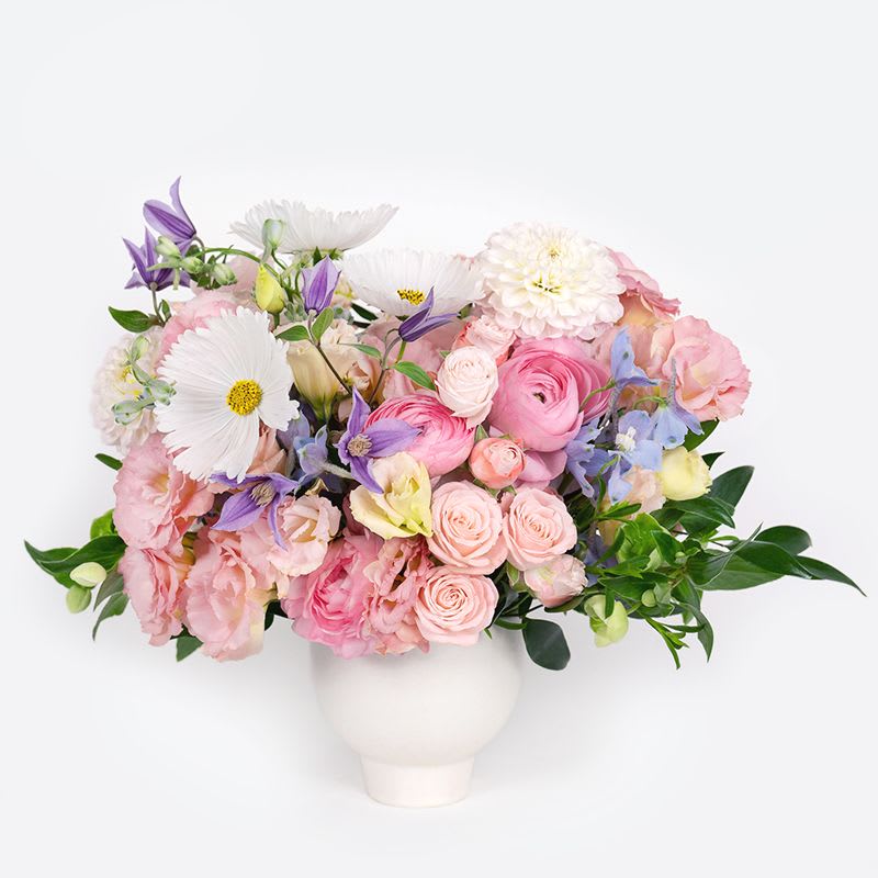Blushing Garden - Soft and romantic, this pastel arrangement features roses, ranunculus, lisianthus, delphiniums, and seasonal blooms in blush, lavender, light blue  and cream tones, accented with fresh greenery. Designed in an elegant 4" compote vase, it’s a timeless centerpiece that brings gentle beauty to any setting