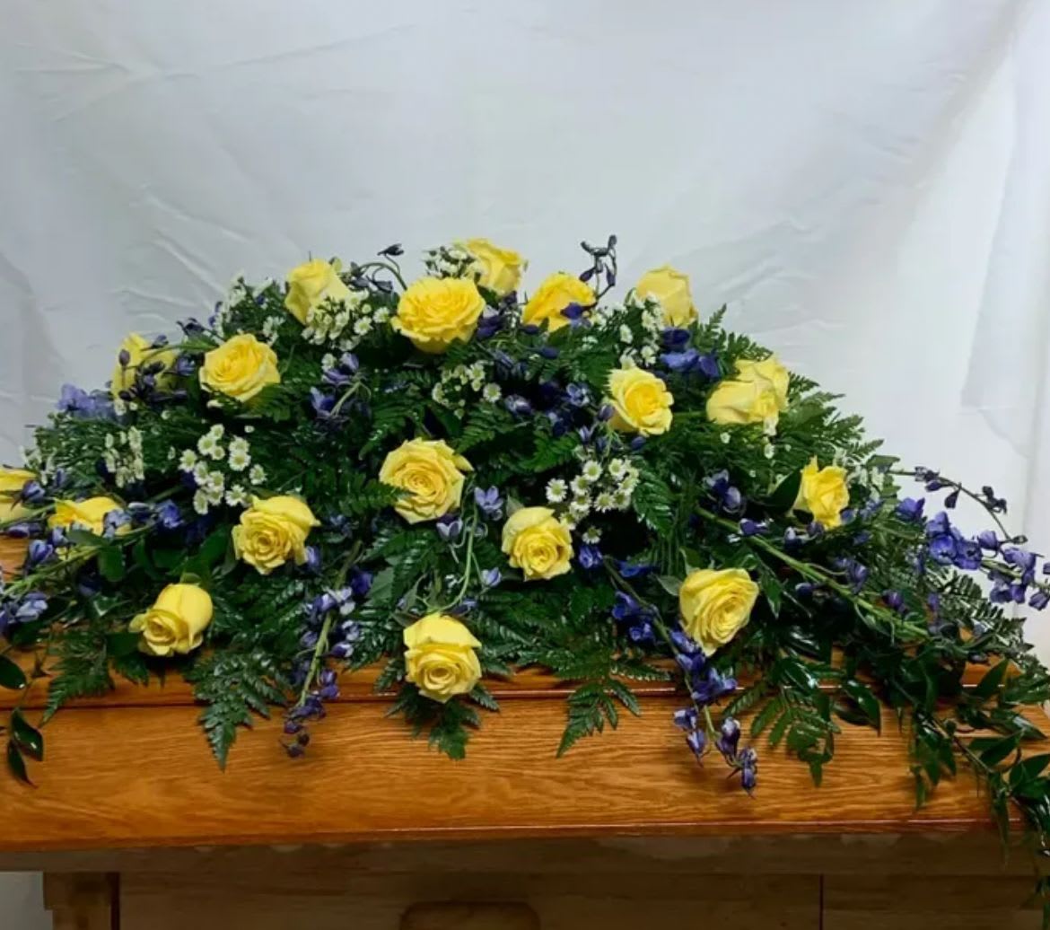 A Shining Life Casket Spray - Bright and uplifting, celebrating a life filled with warmth, joy, and cherishes memories. Radiant yellow roses symbolize friendship, gratitude, and the light that a loved one brought into the lives of others., while striking blue adds graceful height and a sense of peace and admiration.  