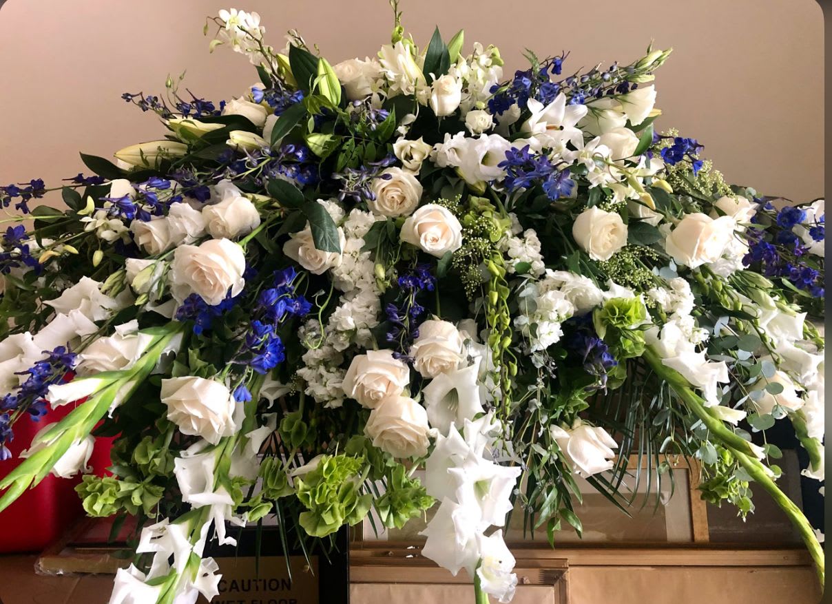 Never Forgotten Casket Spray - A heartfelt tribute to a life that will always be remembered. This harmonious blend of soft ivory blooms and striking blue accents creates a serene and dignified expression of remembrance.  