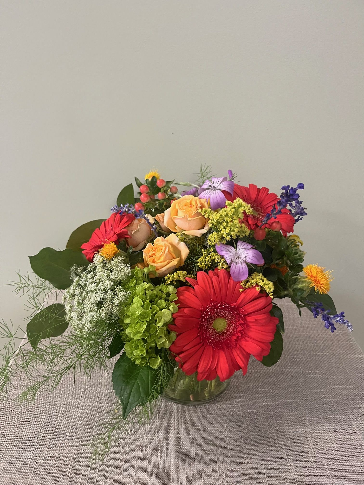 Mixed bouquet with red gerbera daisies, peach roses, and purple flowers in a glass vase