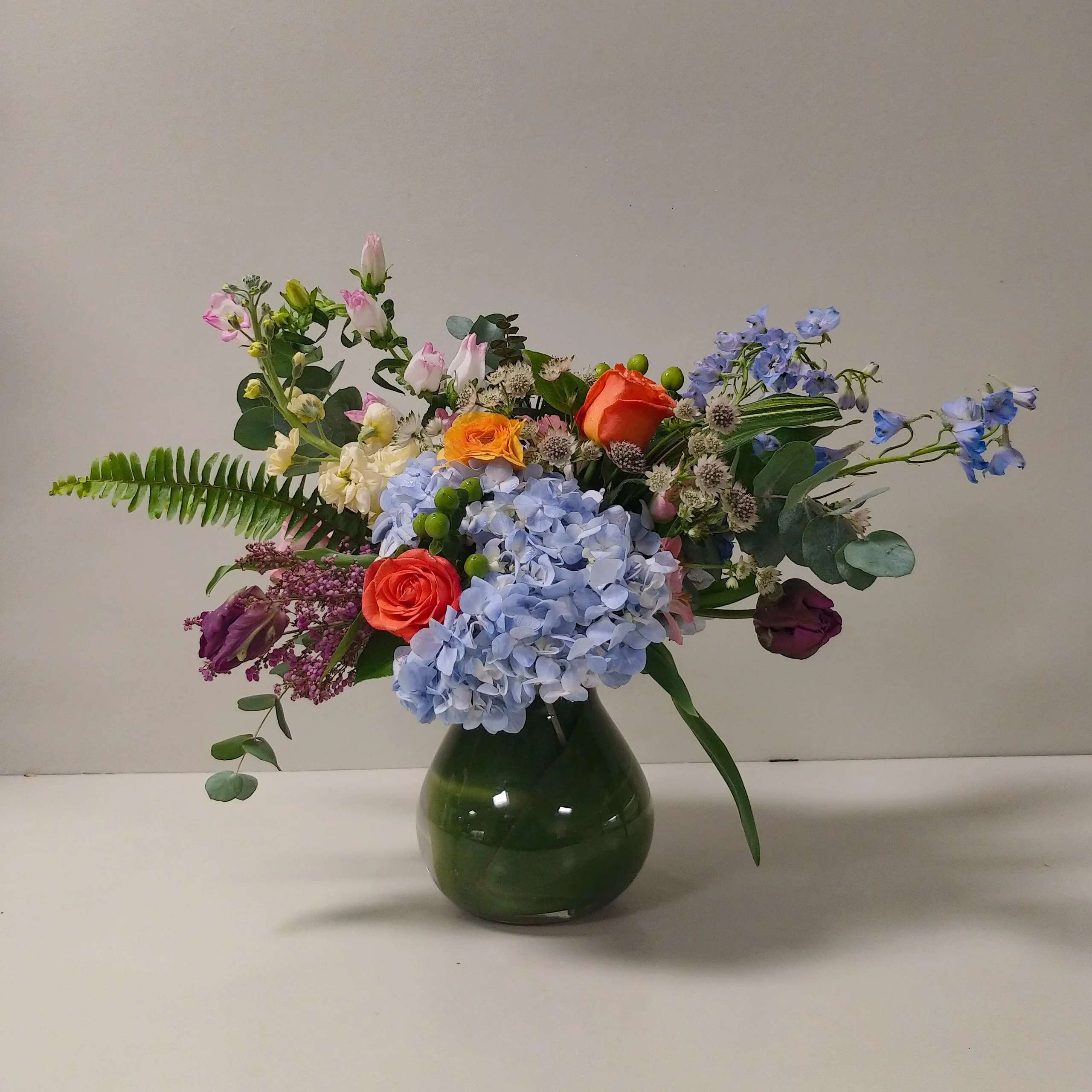 Mixed bouquet in a green glass vase with blue hydrangeas and orange roses