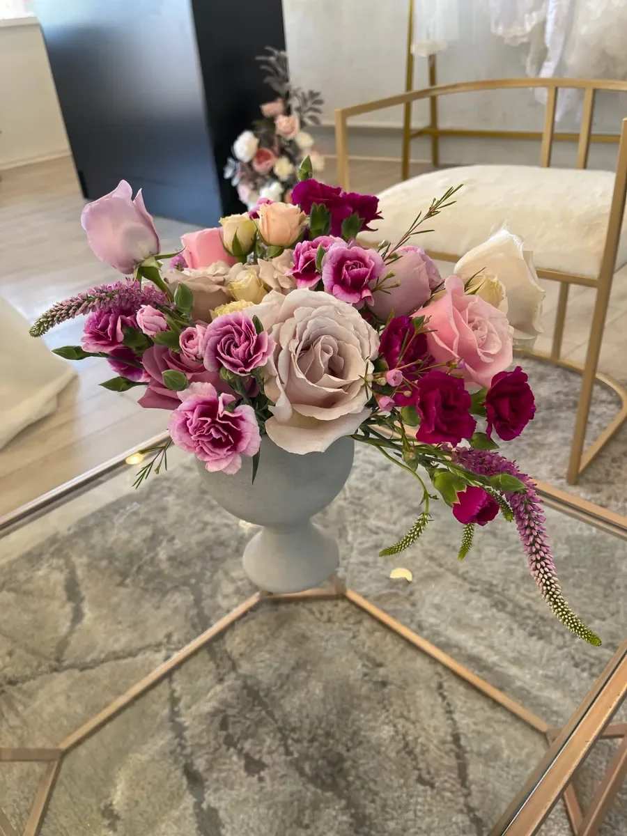 Lilac Passion - A rounded cluster of blush, cream, and deep-pink roses paired with ruffled pink carnations creates a romantic, feminine flower arrangement perfect for a birthday bouquet, anniversary gift, Mother's Day bouquet, or a thoughtful thinking-of-you gesture. The Lilac Passion arrangement reads modern and soft at once-its mixed pinks and purples feel celebratory without being loud, so the person you're sending it to will feel genuinely seen and cherished.  This mixed-color design features roses, carnations, and slender filler spikes arranged compactly in a matte gray pedestal vase that gives the composition contemporary presence on a mantel, bedside table, or entry console. The texture of the ruffled carnations contrasts with the smooth rose petals, while the subtle filler spikes add airiness and movement. The vase provides a stable, finished presentation so the recipient can enjoy the flowers right away.  Ideal for anyone who likes romantic, elegant florals-moms, partners, close friends-Lilac Passion is meant to convey warmth and affection on everyday celebrations or special moments. Order with standard sizing (the arrangement arrives ready-to-display in the pedestal vase); care is simple: keep in fresh water, away from direct heat, and trim stems if you refresh the water to extend bloom life.