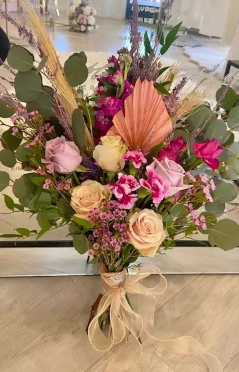 Boho Spring - Cream and blush roses mingle with bright pink blooms and delicate pink filler in this Boho Spring flower arrangement, a romantic bouquet perfect for Mother's Day, birthdays, anniversaries, or a spontaneous "just because" surprise. The recipient will notice the layered, feminine palette-soft pastels warmed by vivid magenta-that reads both fresh and slightly bohemian, a thoughtful way to say thank you or celebrate love and romance.  The hand-tied design is framed with silver-green eucalyptus and accented with dried grasses plus a peach-toned fan-shaped palm for unexpected texture and height. Alongside roses and carnations, smaller filler flowers soften the silhouette and add movement, while a coordinating ribbon keeps the stems elegantly bound. This mix of fresh and dried elements gives the bouquet a tactile, artisanal feel that holds up on a table or bedside.  Give this to someone who should feel seen-mom, a longtime partner, or a friend marking a milestone-and watch it brighten their day. Typically arranged to sit comfortably in a standard vase, treat the fresh flowers with fresh water and routine care (trim stems and change water every few days) to extend their life.