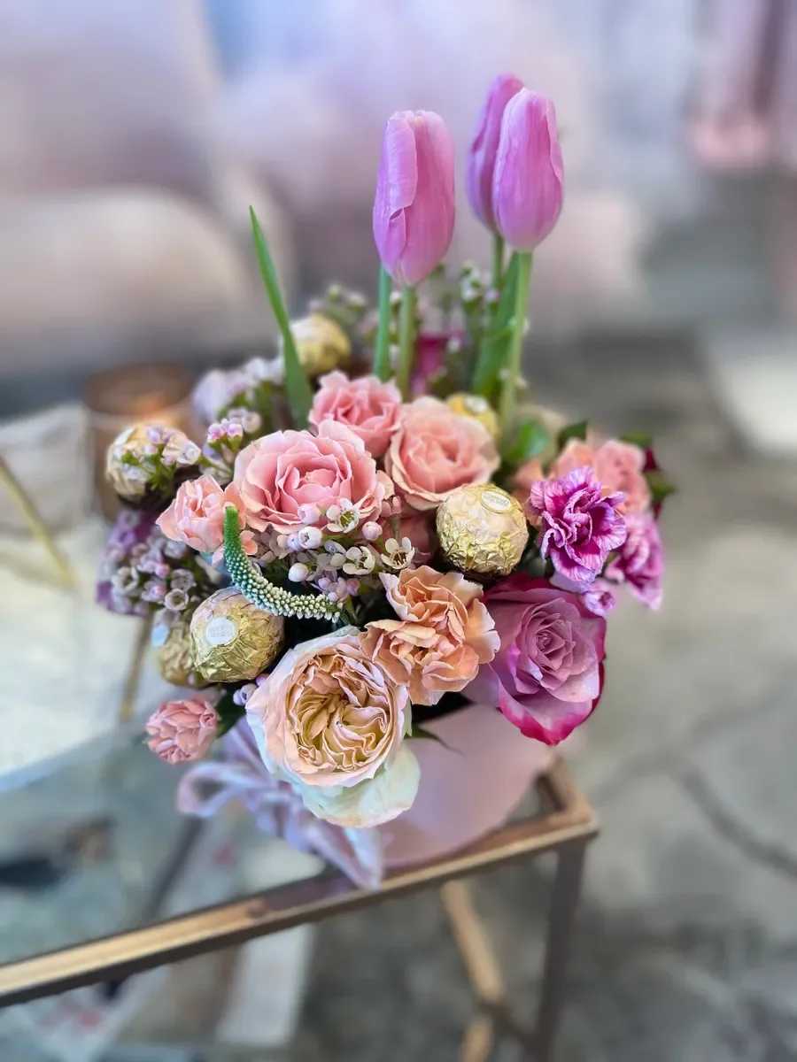 Pink Candy - Soft pink and lavender roses mingle with upright pink tulips in this Pink Candy flower arrangement, an everyday gift bouquet perfect for birthdays, anniversaries, or a sweet just‑because surprise. The pastel palette-pink, purple, white, and hints of peach-creates a romantic, airy impression that will make the person you're thinking of feel cherished and lightly celebrated.  This compact but full arrangement pairs roses, tulips, carnations, and small filler blooms for layered texture and gentle movement. Peach-toned accents lift the composition while a pale pink vase keeps the look soft and cohesive; a white ribbon finishes the presentation. Tucked into the blooms are gold foil‑wrapped chocolates for an extra touch of indulgence, making the gift both beautiful and delicious.  Designed to feel both thoughtful and effortless, Pink Candy suits a close friend, partner, or family member who appreciates pretty pastels and a sweet surprise. The arrangement arrives ready to display in its vase-no fussing required-so she'll feel genuinely seen the moment it's set on her table.