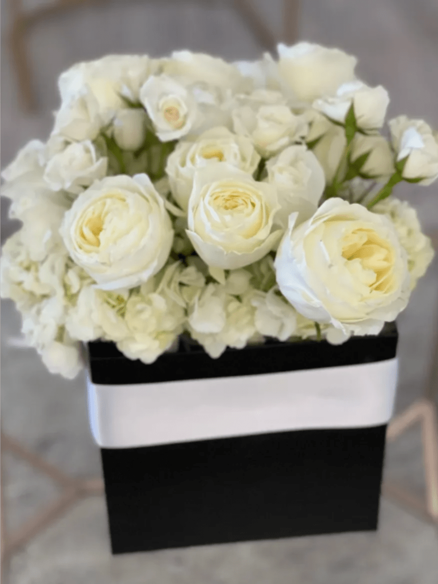 Dior Luxe - Creamy white roses set above a cloud of white hydrangeas create a soft, rounded silhouette in this elegant flower arrangement - perfect as a wedding bouquet alternative, anniversary gift, sympathy tribute, or a refined everyday surprise. The black gift box and wide white ribbon band give the composition a crisp monochrome contrast that reads both romantic and impeccably polished, so the person you're thinking of will feel truly seen and comforted by the composition's quiet luxury.  The arrangement pairs velvety roses and lush hydrangea heads in shades of clean white, arranged densely for a full, sculpted look. Flowers are presented low in a matte black box wrapped with a wide white band for a striking visual and easy tabletop display; the compact, rounded shape makes a bold statement without taking up much space. This is an ideal choice when you want a timeless, formal expression-order in the standard size shown for immediate delivery and enjoy simple care: keep the water topped up and away from direct heat to preserve the blooms.
