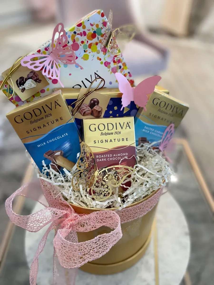 Candy Lux - Candy Lux is a playful gift basket perfect for birthday celebrations or a sweet "thinking of you" surprise. Framing this as a gift basket for casual gifting occasions, it reads like a joyful treat that will make the person you're thinking of feel celebrated and a little spoiled-ideal for a friend, coworker, or someone with a sweet tooth.  Inside a round gold-toned container, several Godiva chocolate bars stand upright amid white shredded paper filler, accented with pink butterfly cutouts and a wide pink ribbon for a feminine, cheerful presentation. The color palette of pink, yellow, blue, and white comes through in the bright packaging and delicate embellishments, and included gift tags make adding a personal message easy.  This is the kind of ready-to-give set that works for informal moments-thank-yous, congratulations, or just because. Arrange for local delivery or pickup to ensure the ribbons and butterflies arrive neat; no special care required beyond keeping chocolates in a cool place.
