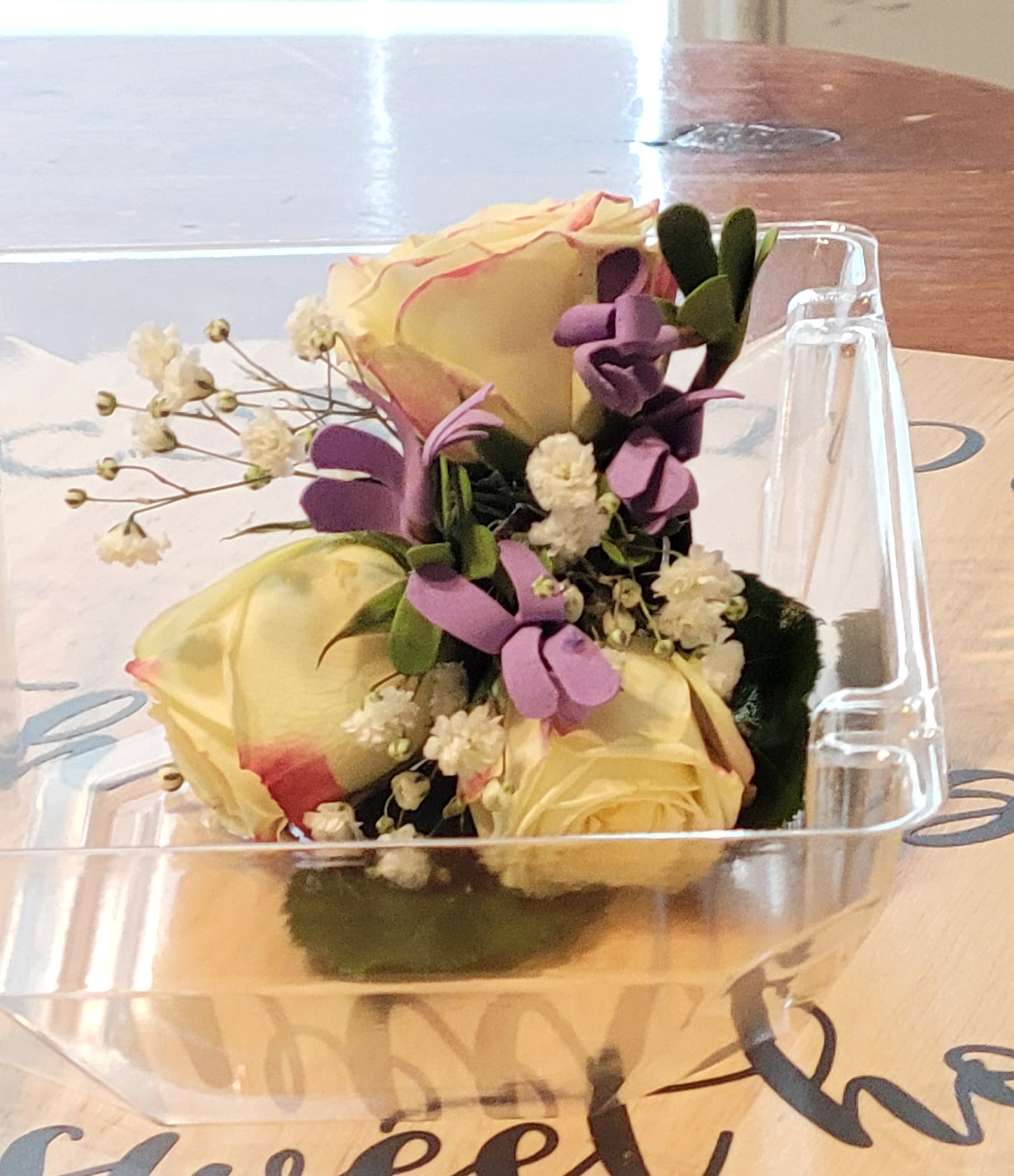 Small bouquet of pale yellow roses with purple accents in a clear plastic box