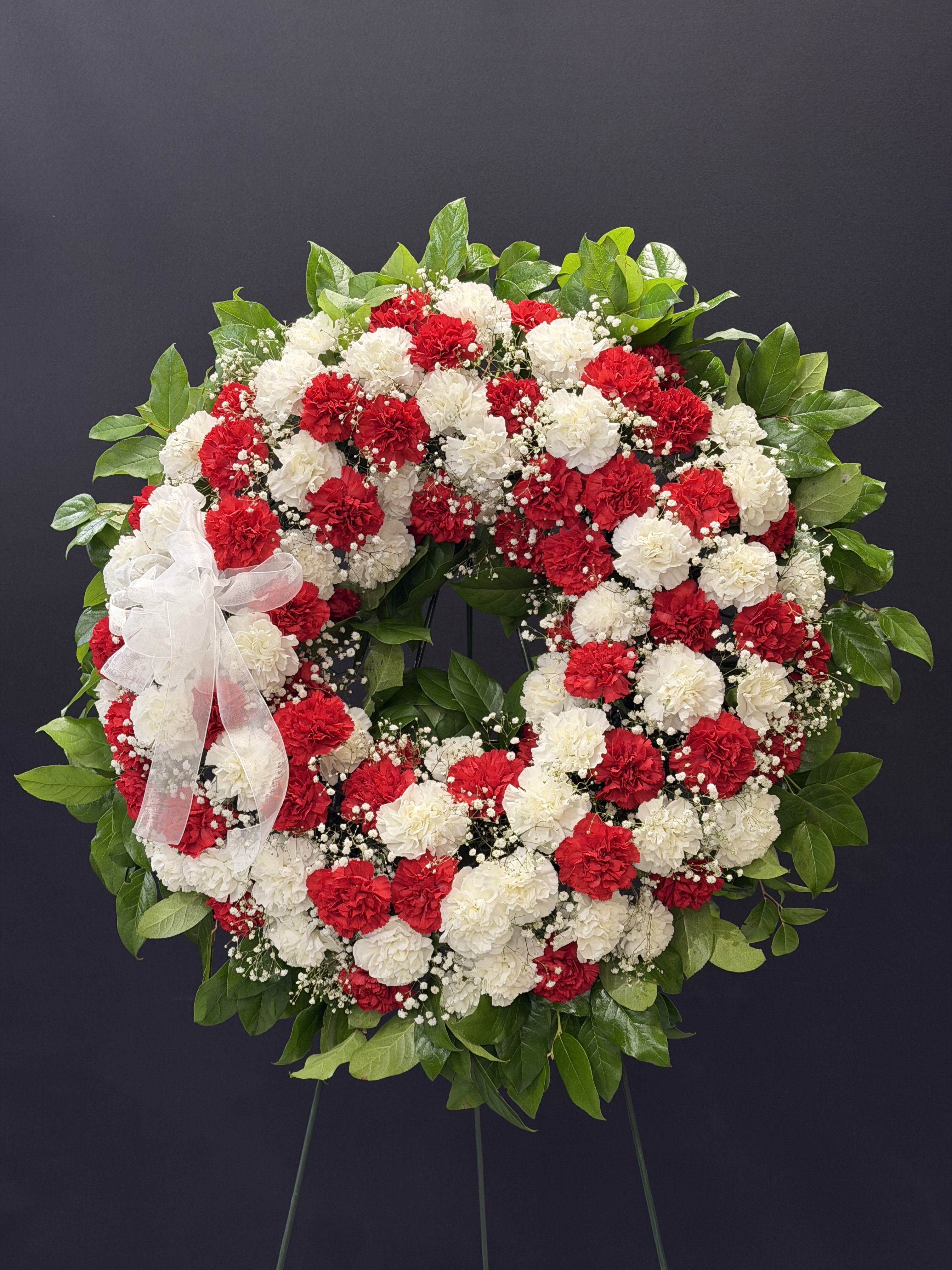 Circle of life - A large circular sympathy wreath crafted from red and white carnations creates a dignified, heartfelt tribute for a funeral or memorial service. The bold red blooms paired with pure white carnations and delicate sprays of baby's breath deliver a respectful, ceremonial impression-the kind of arrangement that helps the person you're honoring feel remembered and cherished.  Glossy green foliage frames the outer edge and fills the center opening for a full, layered silhouette, while a sheer white ribbon bow on the left side adds a soft, formal accent. Mounted on a wreath stand and built on a sturdy wreath form, this arrangement arrives ready for display and makes a lasting visual statement at services or at a graveside.