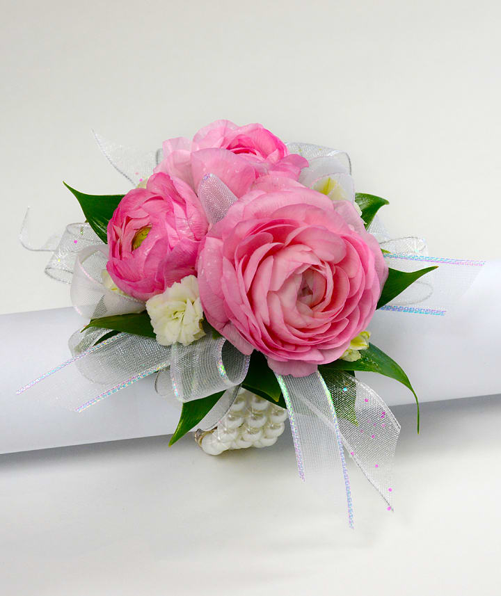 Pink ranunculus wrist corsage with white accent flowers and sheer ribbon on a pearl bracelet.