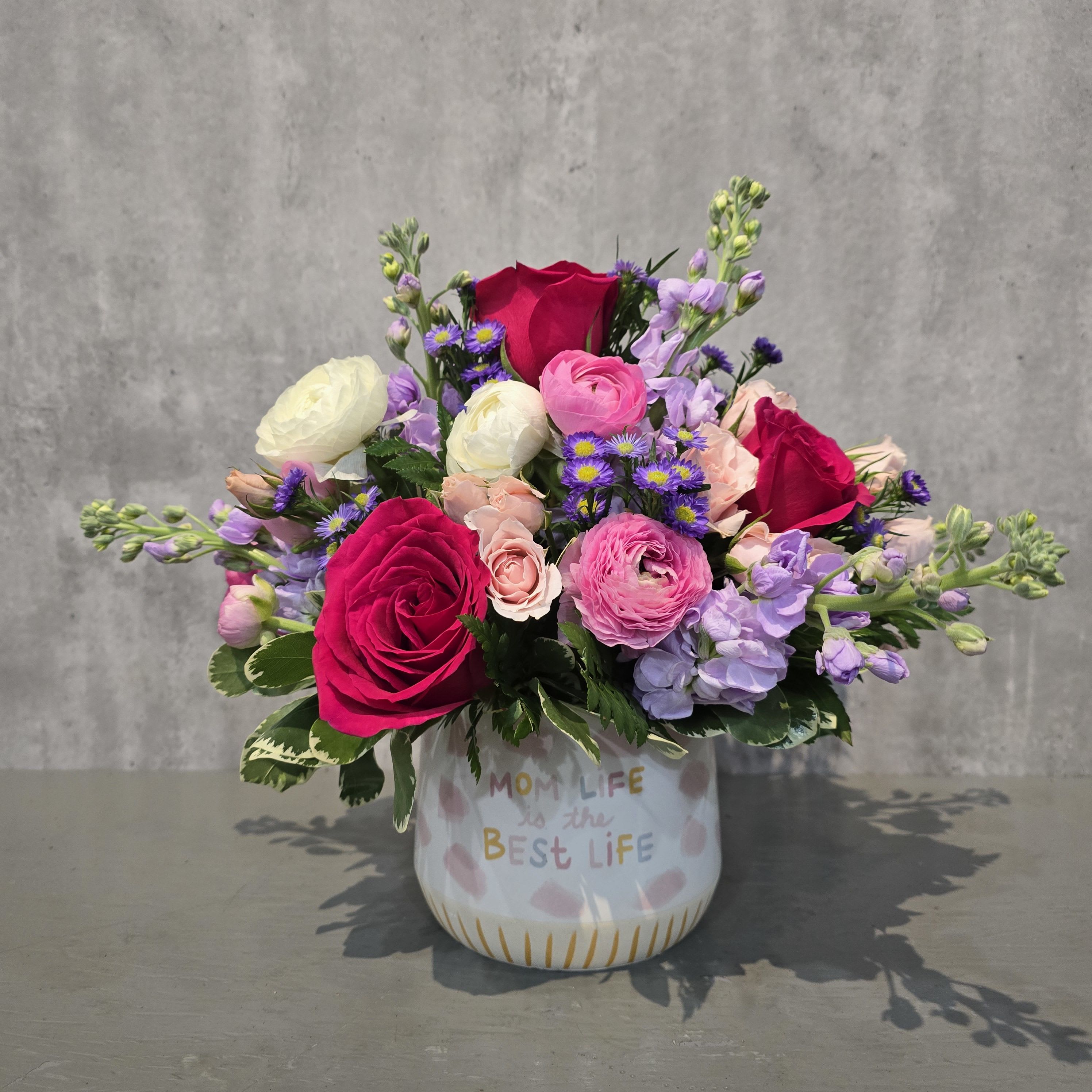 Best Life Blooms - Celebrate everything Mom is with Best Life Blooms, a cheerful floral arrangement designed in a keepsake container that reads “Mom Life is the Best Life.” Bursting with fuchsia roses, white and pink ranunculus, soft peach spray roses, lavender stock, and purple asters, this compact design is full of heart and color. Measuring approximately 12" tall and 13" wide, it's the perfect size for desks, bedside tables, or kitchen counters. This handmade arrangement is a sweet way to show appreciation and is available for Mother’s Day delivery throughout the area.