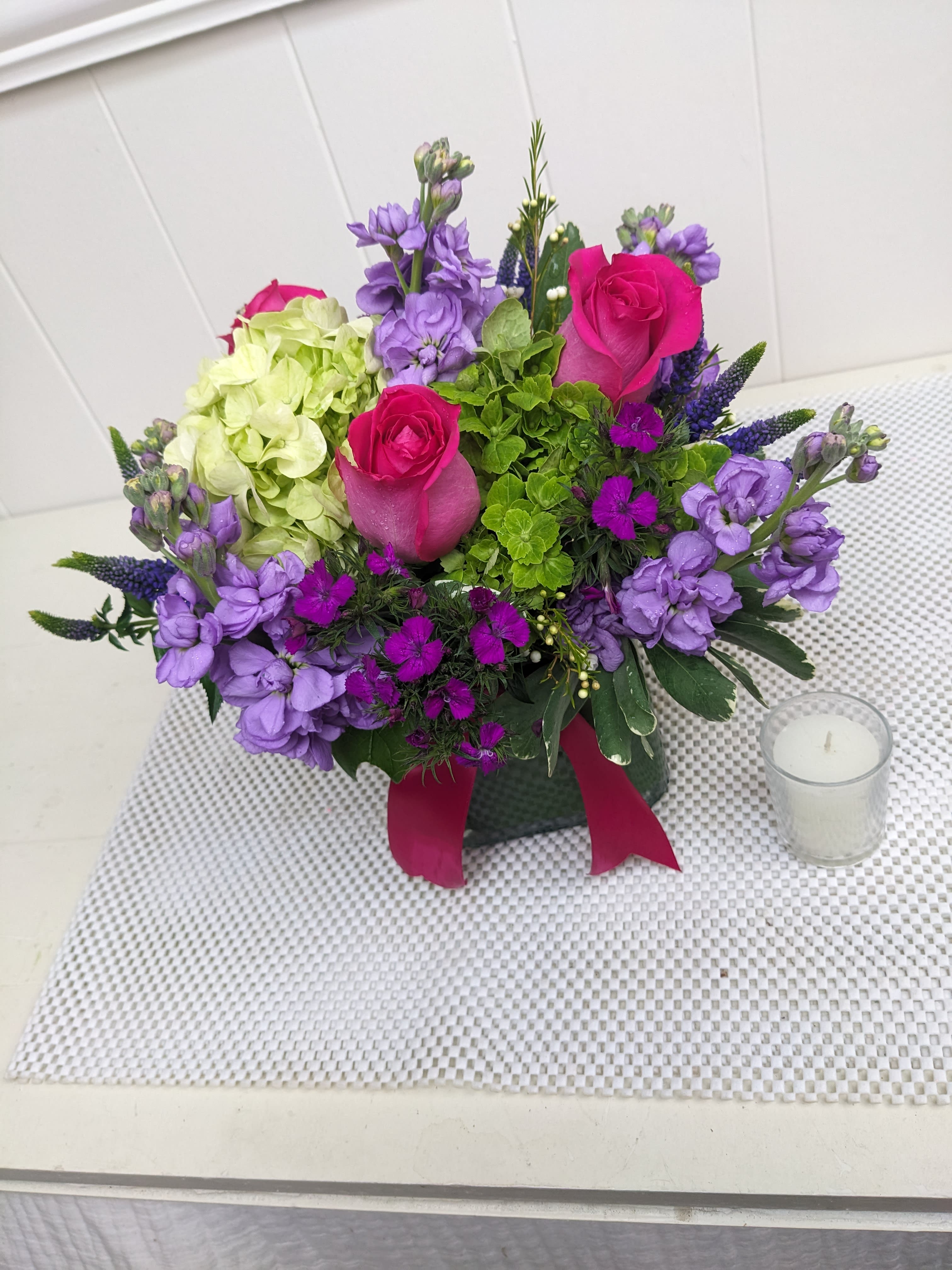 Compact arrangement of hot pink roses, green hydrangea, and purple blooms in a glass vase with ribbon
