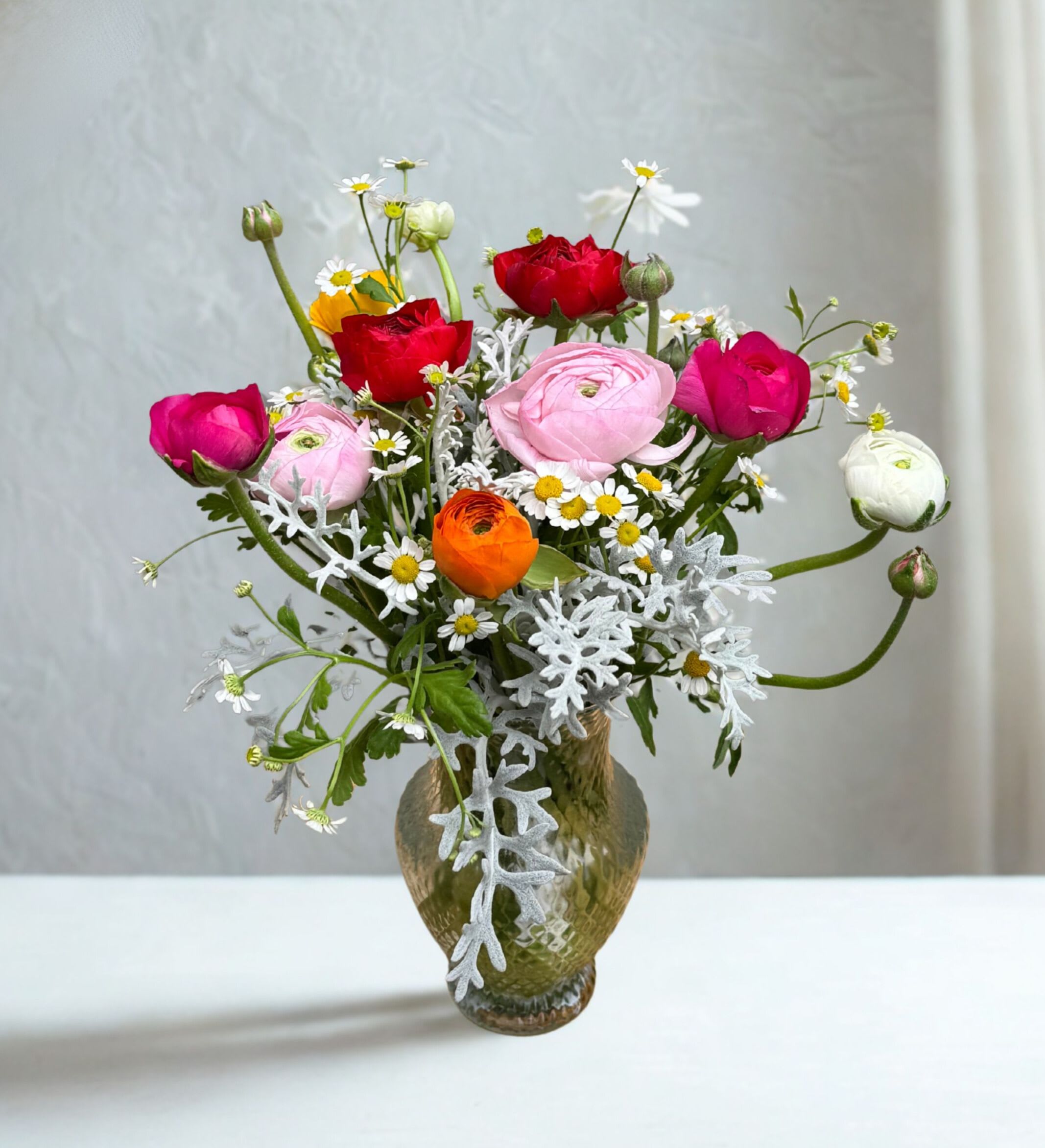 Tall mixed bouquet of pink, red, white, and orange ranunculus with small daisies in a green glass vase