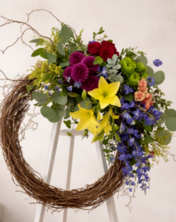 Funeral arrangement on the easel with natural elements  - A bold circular wreath of mixed-color blooms mounted on a white easel-this funeral arrangement on the easel with natural elements offers a respectful, yet celebratory tribute for a memorial service or ceremony. The grapevine wreath base gives the piece an organic, rustic character while the saturated palette of deep red roses, bright yellow lilies and purple chrysanthemum-like blooms reads clearly from a distance, so the person you're honoring will stand out among the floral displays.  Up close the composition is richly layered: clusters of red roses and yellow lilies sit beside green pom‑pom chrysanthemums, peach accents, and trailing blue delphiniums sprays woven through airy eucalyptus and complementary foliage.   Choose this piece when you want a meaningful, stand-alone tribute that feels both heartfelt and visually striking-the kind of arrangement that helps mourners remember and celebrate. For delivery and placement, note that the wreath is mounted on the easel and stand for easy setup at the venue; contact us if you need assistance with timing or on-site placement.