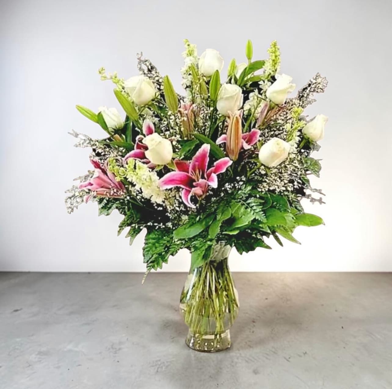 One Dozen White Roses with a Flair - This stunning arrangement of a dozen White Rose is accented with Star Gazier Lilies, Larksburg and filler with  3 types of Greenery designed in a clear glass vase. 