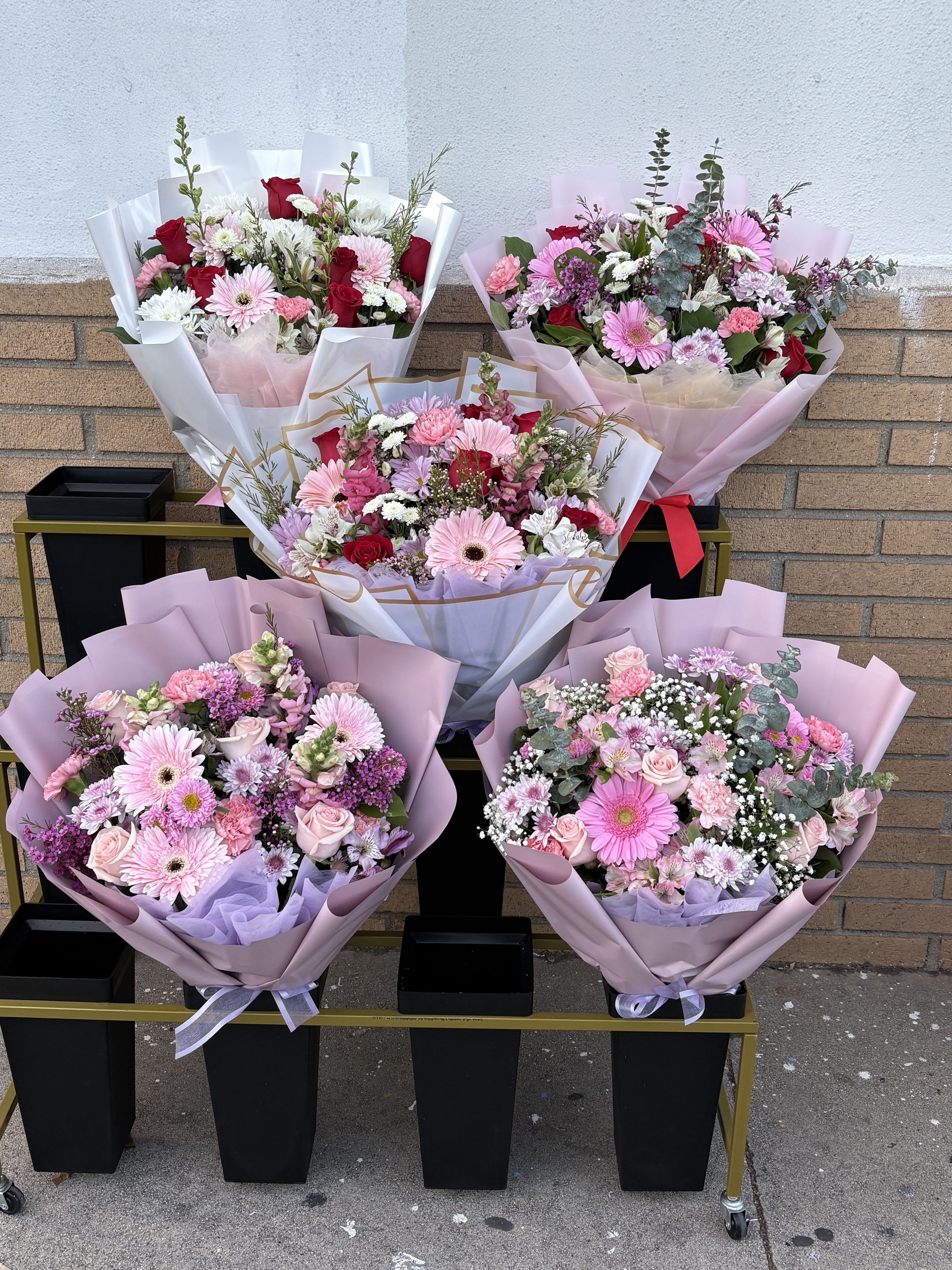 Five pastel flower bouquets arranged on a cart against a brick wall