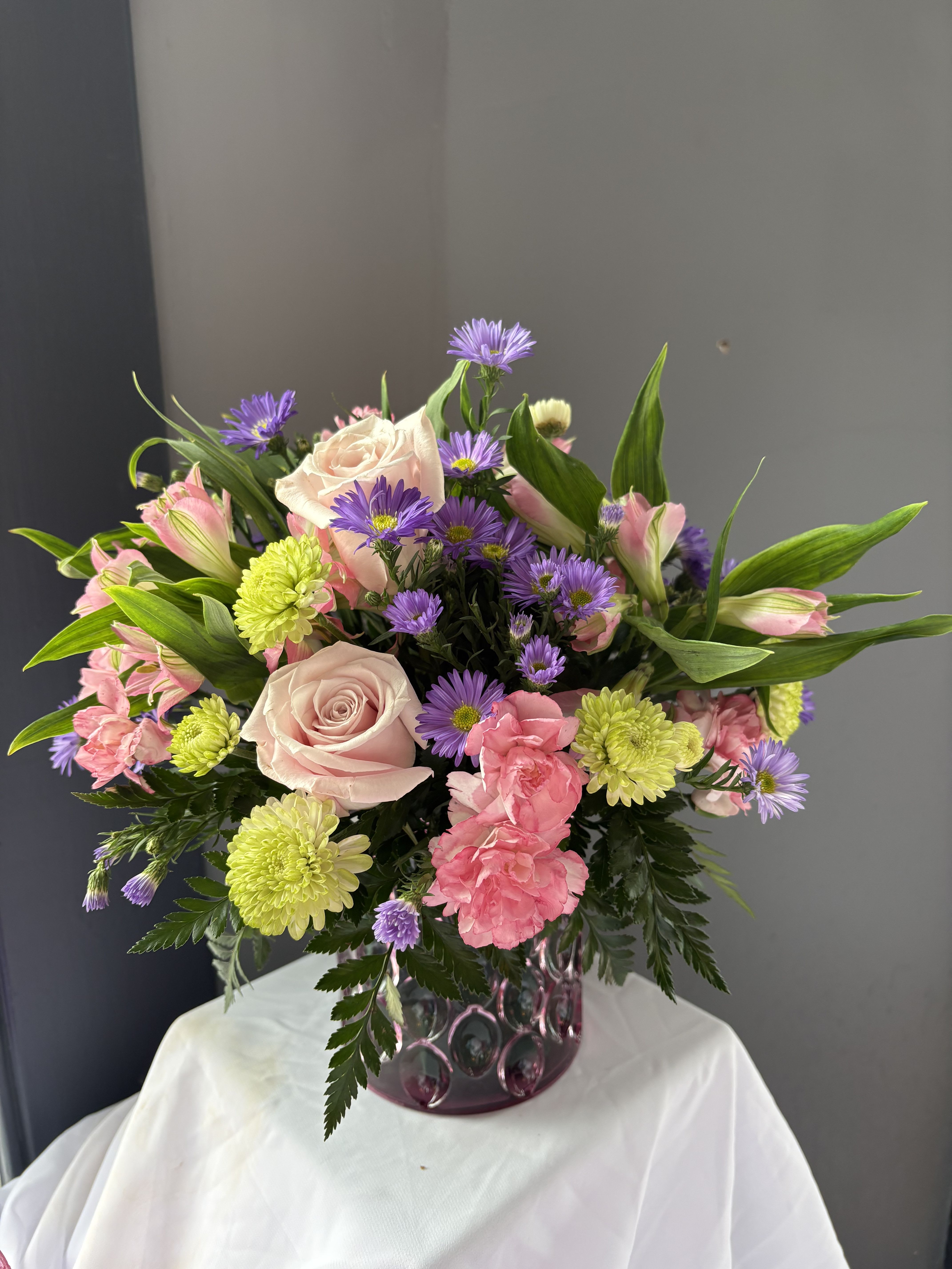 Frazier's Verison Pink Rhapsody Bouquet - Celebrate her in style with Frazier's Version Pink Rhapsody Bouquet, a vibrant mix of pink roses, green buttons, pink mini carnations, and purple monte beautifully arranged in Teleflora's Pink Rhapsody Vase. This stunning Mother's Day gift adds bold color and lasting charm to any space. The scale of the arrangement should determine the type of substitution acceptable, but the substitution must be of equal or greater value and must maintain the style and color harmony of the original order.