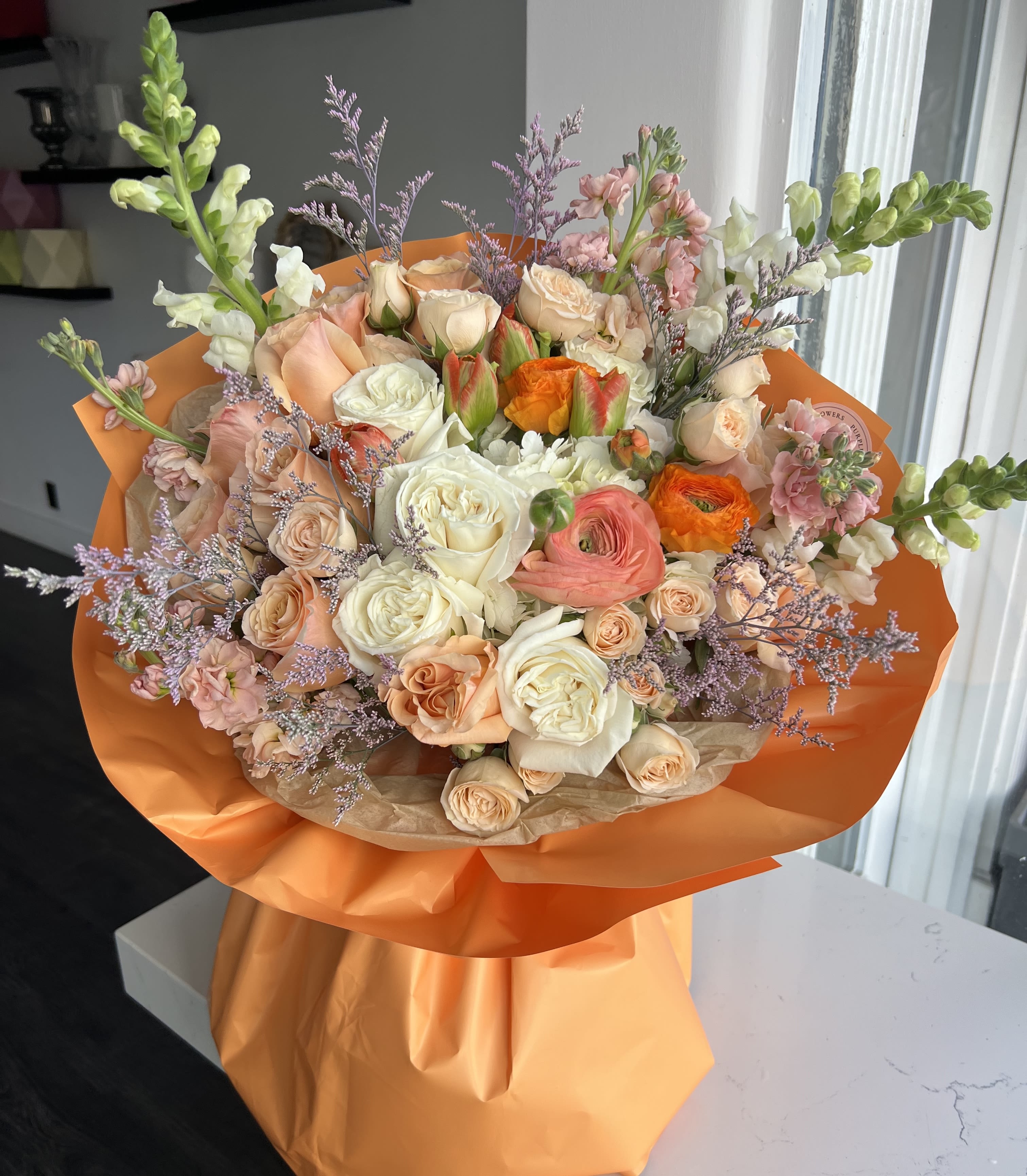 Hand-tied bouquet of peach and white roses with orange ranunculus wrapped in bright orange paper.