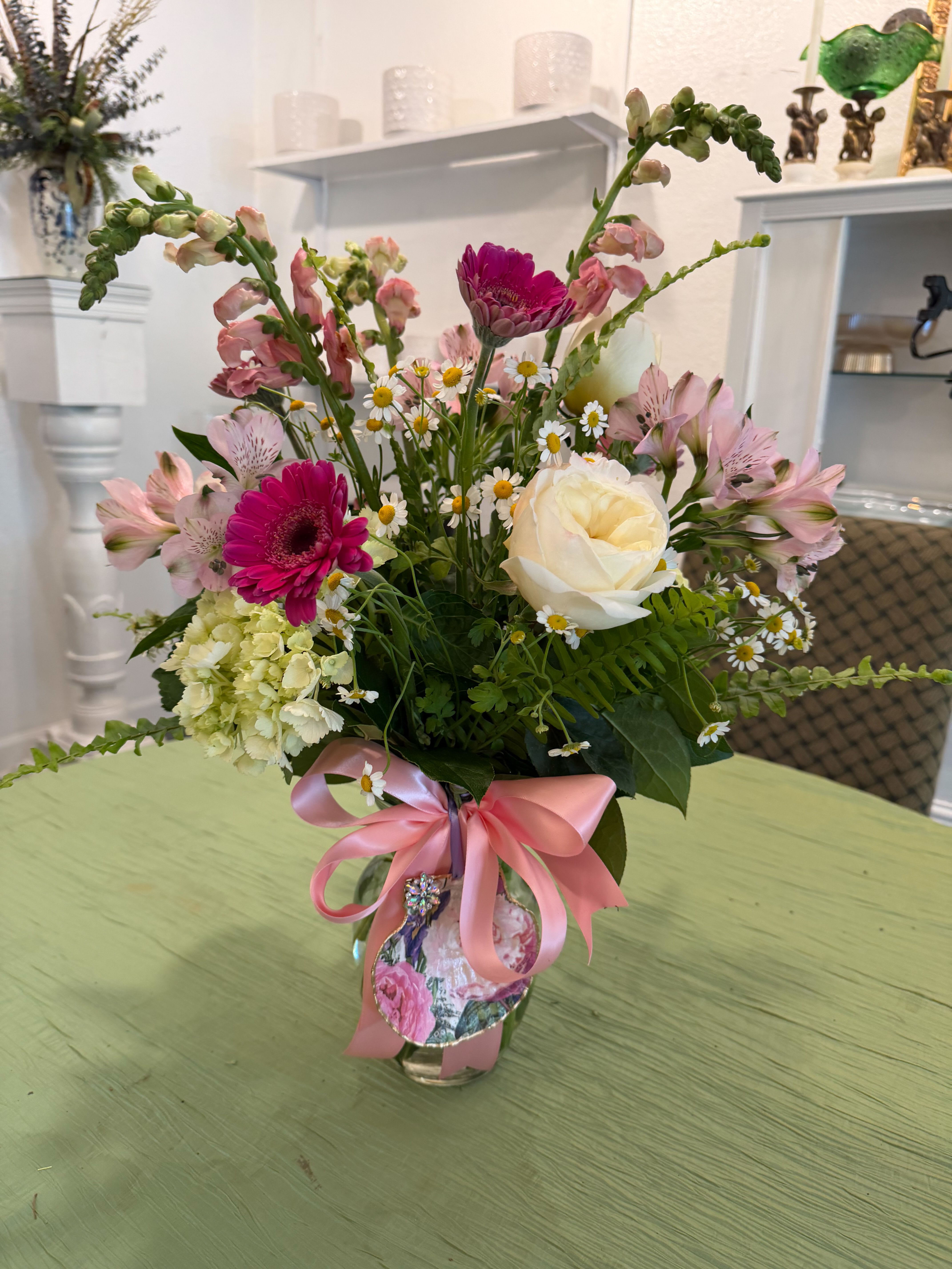 Mother's Day Mix - The Mother's Day Blooms flower arrangement, a cheerful  Mother's Day gift that reads like a fresh-picked garden. This spring mix is perfect for celebrating mom, a grandmother, or any woman who lights up a room.  The mixed pink, white and green palette feels feminine and uplifting without being fussy. Arranged in a clear glass vase and finished with a pink satin ribbon and decorative decoupaged shell, it arrives gift-ready and easy to display on a kitchen counter or bedside table.   Choose this arrangement to mark Mother's Day, a birthday, or to say "thinking of you" with something that will make her feel truly celebrated. Vase size and stem lengths are arranged for a medium, table-friendly profile; change water every two days and trim stems slightly for best vase life.