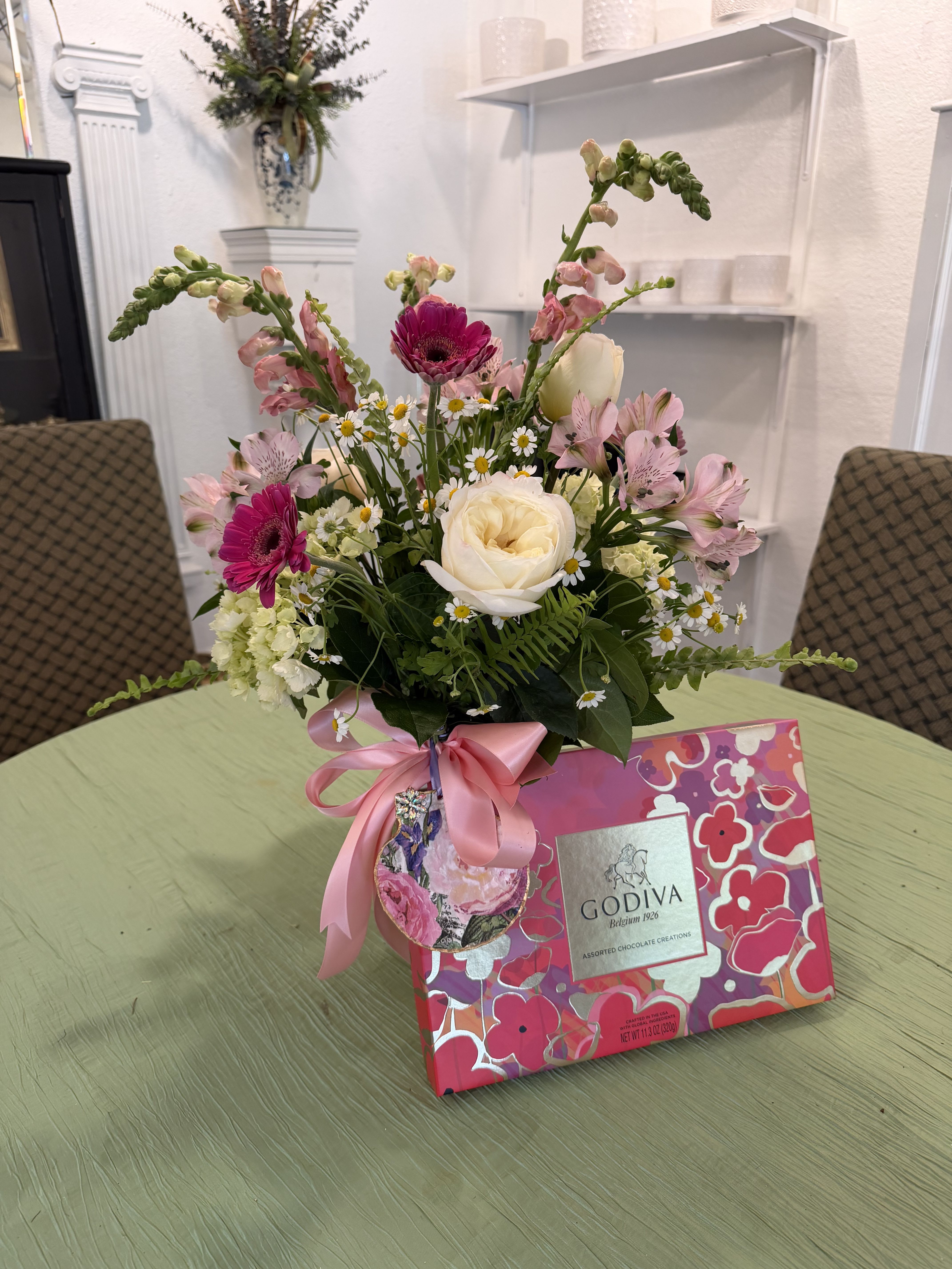 Mother's Day Mix with Chocolates  - The Mother's Day Blooms flower arrangement, a cheerful Mother's Day gift that reads like a fresh-picked garden. This spring mix is perfect for celebrating mom, a grandmother, or any woman who lights up a room. The addition of chocolates makes it even better!  The mixed pink, white and green palette feels feminine and uplifting without being fussy. Arranged in a clear glass vase and finished with a pink satin ribbon and decorative decoupaged shell, it arrives gift-ready and easy to display on a kitchen counter or bedside table.  Choose this arrangement to mark Mother's Day, a birthday, or to say "thinking of you" with something that will make her feel truly celebrated. Vase size and stem lengths are arranged for a medium, table-friendly profile; change water every two days and trim stems slightly for best vase life.