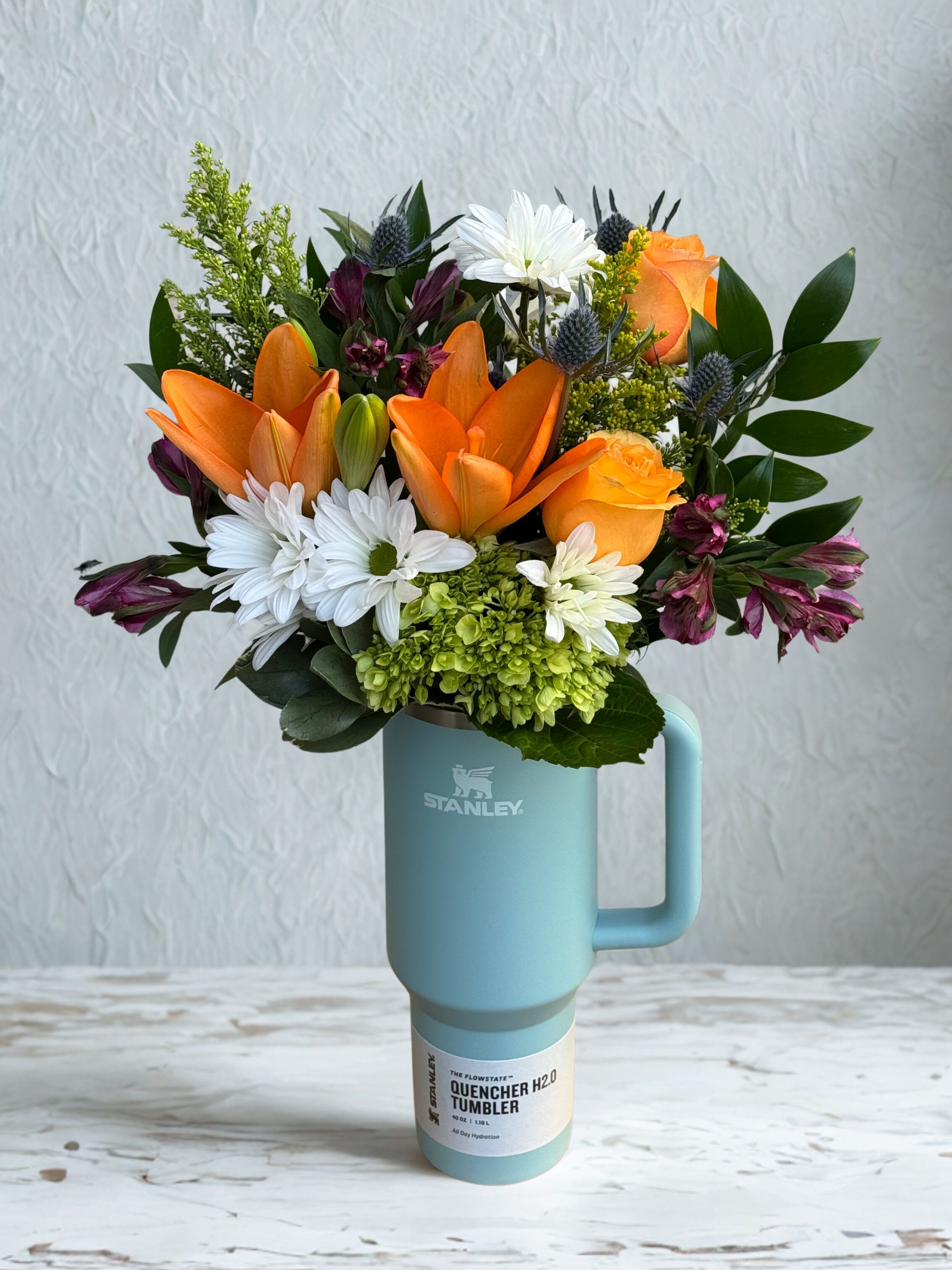 Mom's Morning Mug - The perfect gift for any Stanley lover. Each arrangement features a seasonal mix of flowers, thoughtfully designed by our florists. Images shown represent the different price options, with the deluxe version showcasing more premium blooms.  Limited Quantities Available