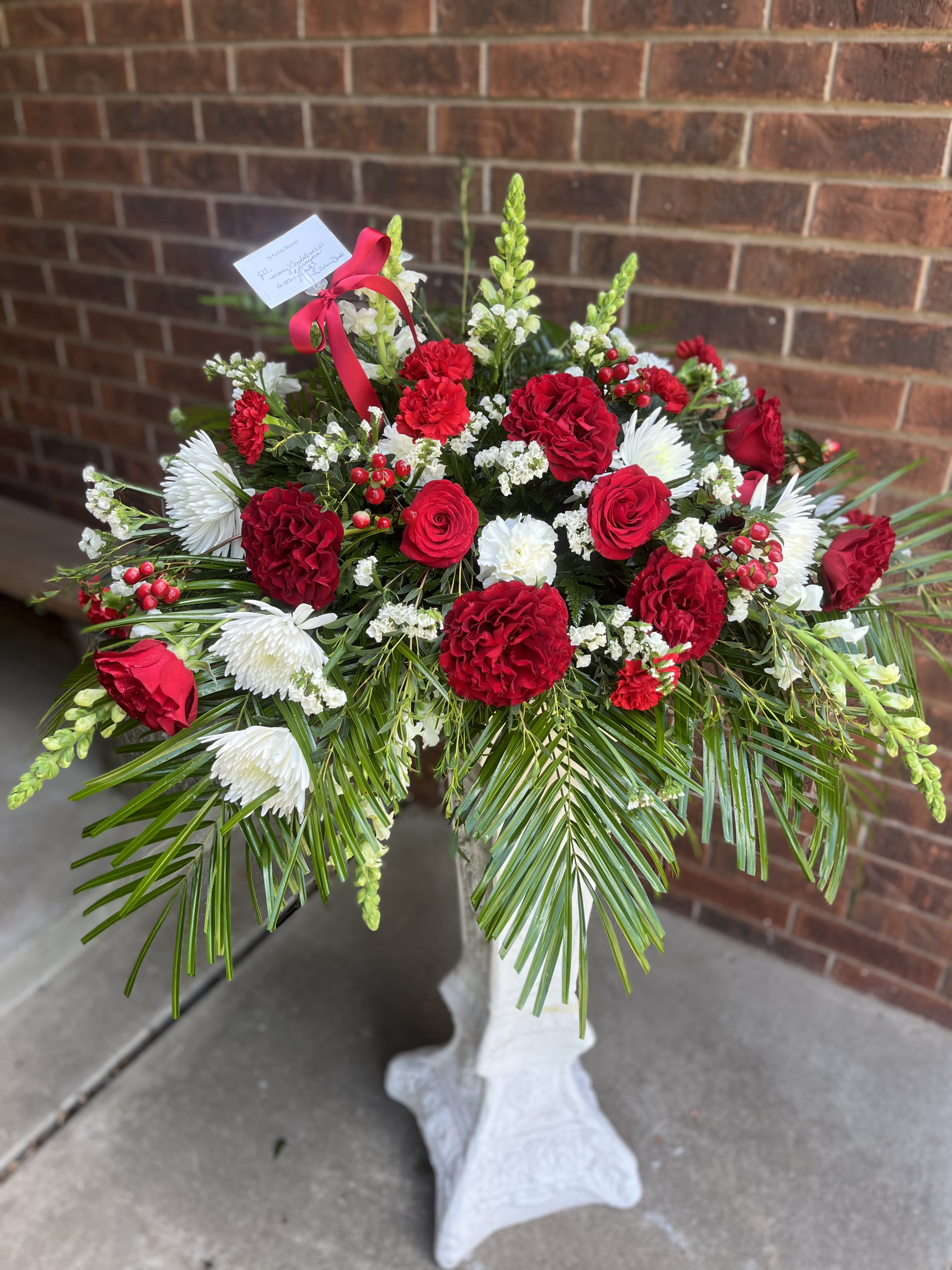 Red Hearts Tribute - A bold spray of red roses and carnations centered against pure white chrysanthemums creates an immediate, reverent impression-perfect for a sympathy arrangement or memorial tribute. This standing funeral spray communicates deep love and respect for the person you're honoring, the kind of floral statement that helps grieving friends and family feel held and remembered.  Dense, rounded clusters of red roses and red carnations form the heart of the design, accented by white chrysanthemum blooms and tall white snapdragon spikes that lift the composition. Long palm fronds and layered greenery fan outward to give the spray a broad, ceremonial silhouette; finished with a red ribbon, a small card, and mounted on a standing easel for display.  Designed to be placed at a service, graveside, or memorial gathering, this traditional standing spray conveys heartfelt sympathy and lasting remembrance. The arrangement arrives ready to display on the included easel; for freshness, keep flowers in a cool, sheltered spot before the service and follow any care instructions provided on the card.