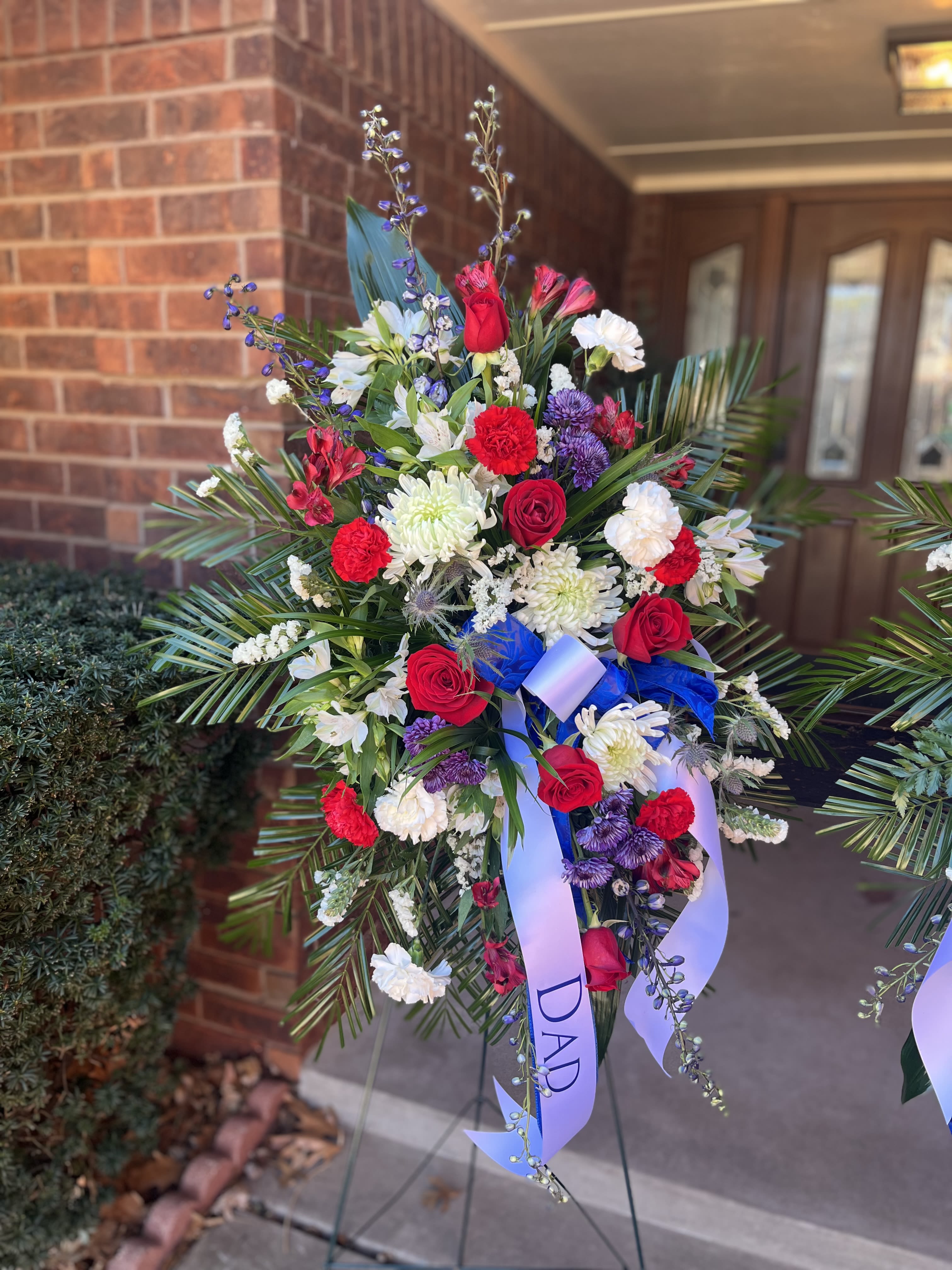 Salute Standing Spray - A bold tribute anchored by deep red roses and crisp white blooms, this standing spray is designed as a sympathy arrangement for a father or beloved family member. The tall, layered silhouette-built on a standing easel-creates a formal, expressive memorial presence that will make the recipient feel honored and remembered.  The spray combines red roses, white chrysanthemums and carnations, blue accents, alstroemeria and delicate filler flowers for texture, with long palm fronds and airy branching stems to add height and movement. Rich color contrast between the reds, whites, purples and subtle blue ribbon gives the piece visual depth from every angle; the arrangement is densely composed at the center and tapers naturally to the edges for a classic standing-spray profile.  Perfect for a funeral service, graveside tribute, or memorial gathering, this piece sends a clear message of respect and love for the person you're honoring. It arrives ready to display on the included easel and is designed to hold its form throughout services; order with any personalization requests for the ribbon to ensure the message reads exactly as you intend.  Ribbon optional