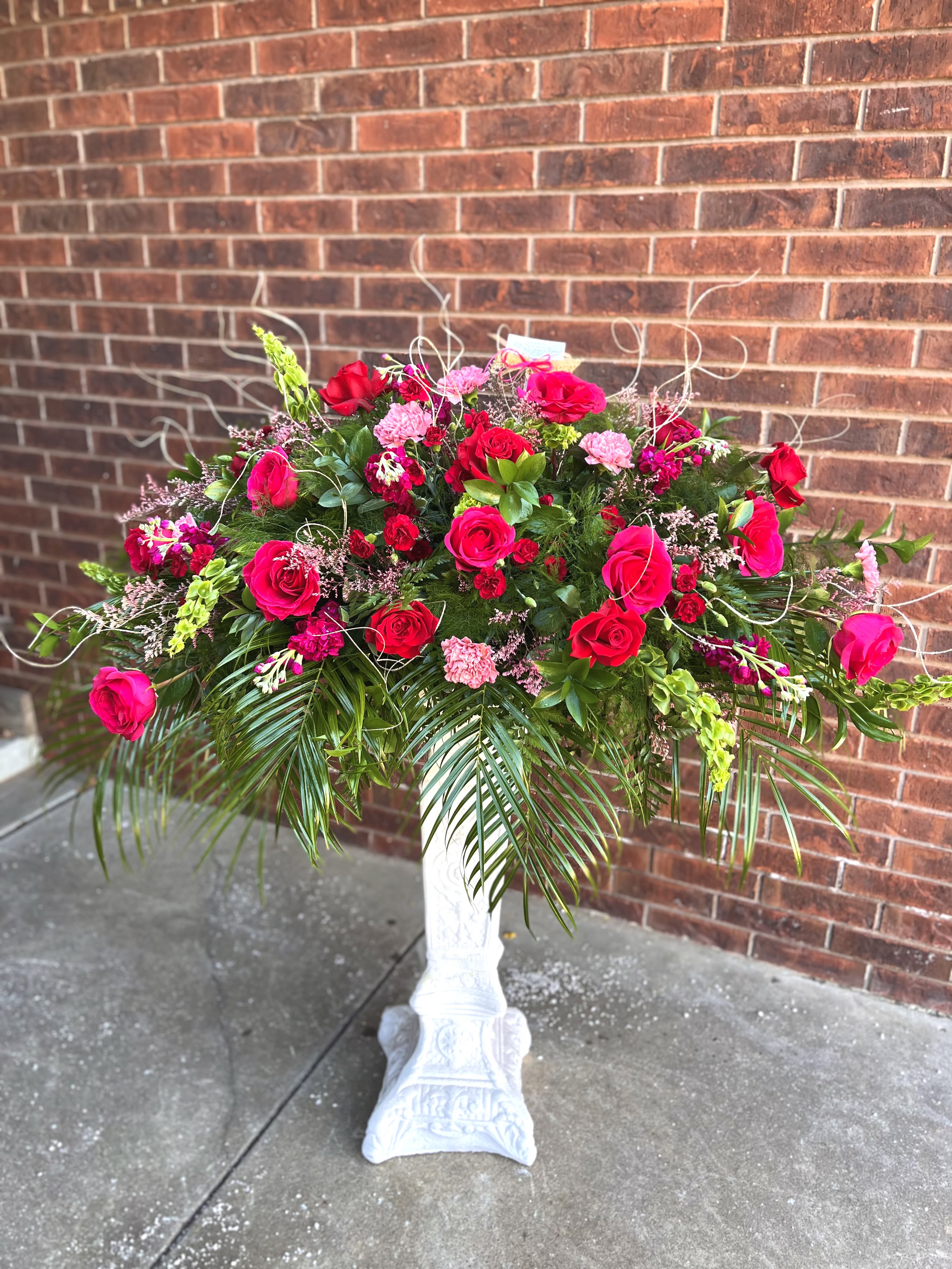 Rosie Palasades - A bold, celebratory display of pink and red florals that makes a statement for a wedding reception, anniversary, or grand opening. This large, sprawling flower arrangement is a dramatic centerpiece-style bouquet perfect as a birthday bouquet for someone who loves vivid color or as an attention-grabbing sympathy or event arrangement when you need to mark a meaningful moment.  Roses and carnations create the lush, romantic core in shades of pink and red, lifted by tall snapdragon spikes and airy filler for height and movement. Long palm fronds and trailing greenery spill over the base for a tropical, sculptural silhouette, while touches of white blooms add bright contrast and visual relief. The horizontal, wide design fills a mantle or ceremony table.  Made to be seen from all sides, this arrangement suits recipients who should feel celebrated and noticed-the kind of gift that reads "this is important." For placement, choose a prominent table or entry display; water as needed and keep out of direct heat to extend freshness.