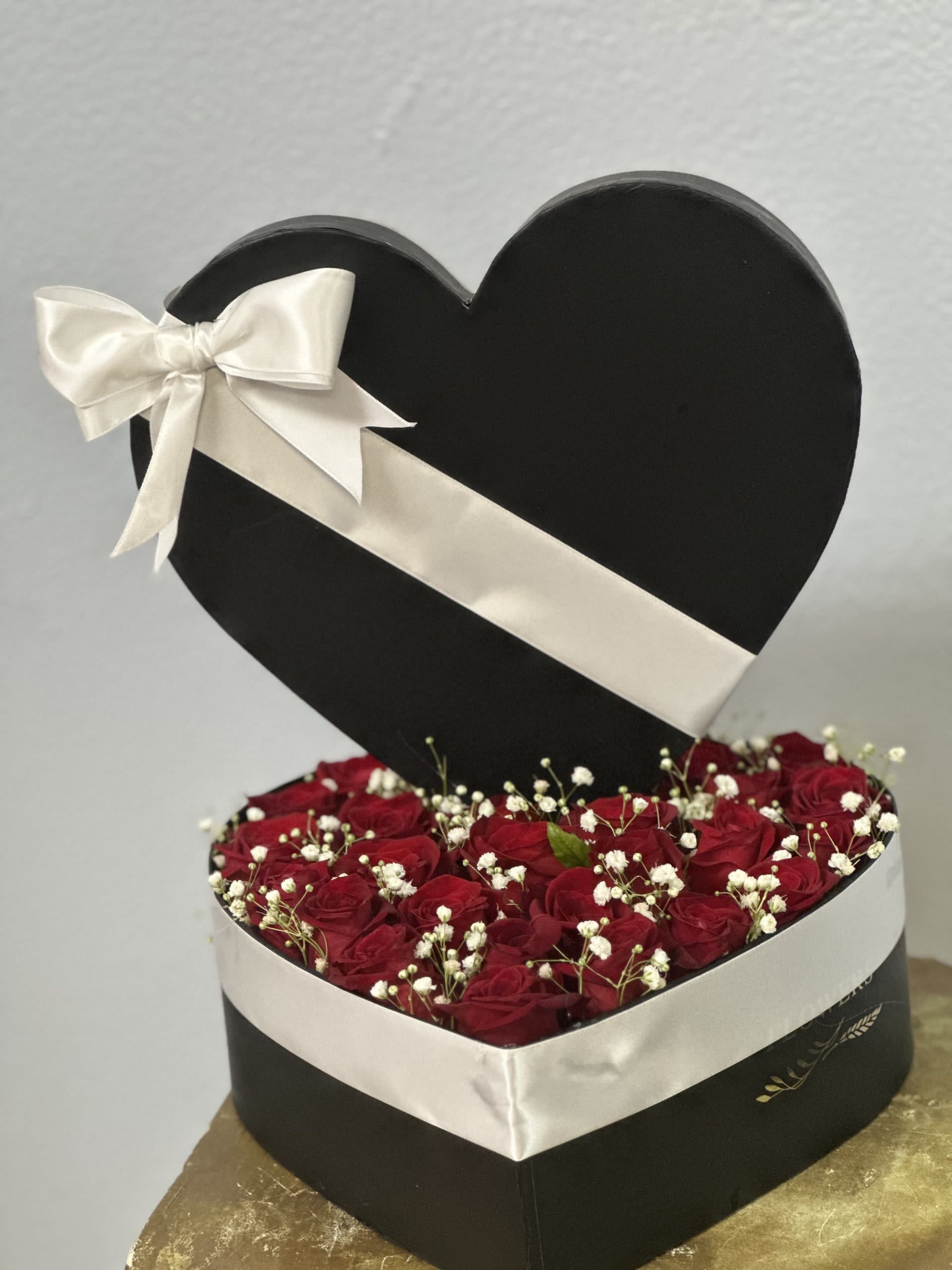 Midnight Heart Rose Box - Deep, romantic, and unmistakably luxe - this heart-shaped flower arrangement makes a bold anniversary or Valentine's Day gift that reads as passion and thoughtfulness in equal measure. The heart-shaped black gift box holds a tightly packed bouquet of deep red roses with delicate white baby's breath scattered through the blooms, creating a striking red, white, and black palette that draws the eye and stirs the heart.  The box is wrapped in a wide white satin ribbon tied into a large bow on the lid for a polished, gift-ready presentation; the compact, low profile keeps blooms close together for a lush, dense look. Sized to sit on a bedside table or mantel, this arrangement is ideal for the person you want to make feel cherished and seen-perfect for anniversaries, romantic gestures, or a dramatic celebration of love. Care is simple: keep in a cool spot away from direct sunlight and check water as directed to keep roses fresh.