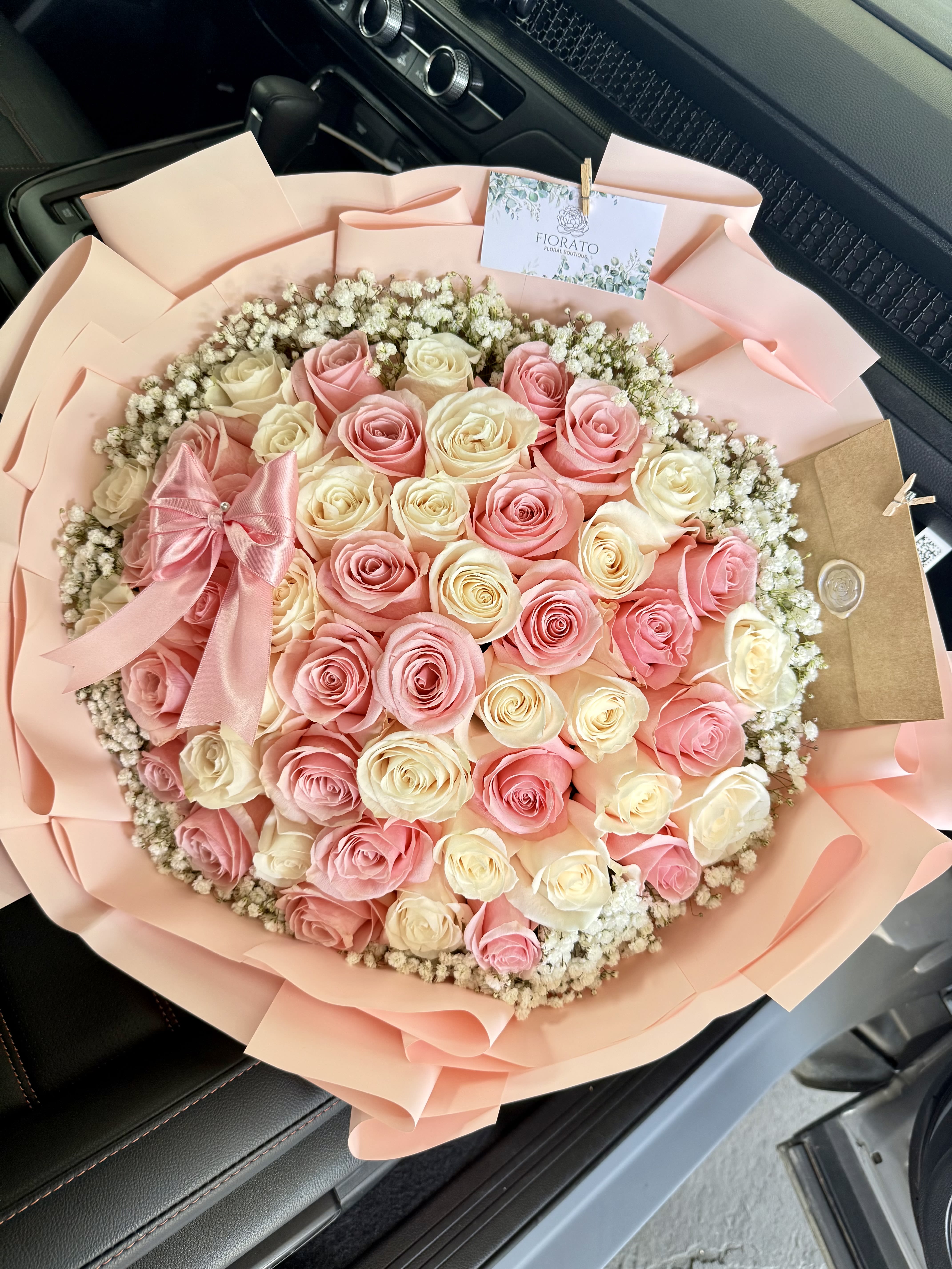 Alejandra - Fiorato Sacramento, CA. - A lush, round bouquet of tightly packed pink and cream roses edged with a halo of tiny white filler flowers - perfect as a birthday bouquet, anniversary roses, or a Mother's Day gift that reads as both celebratory and tender. The soft pastel palette of pink, white, and peach creates a romantic, feminine impression that will make the person you're thinking of feel cherished and seen.  Arranged full and low for maximum visual impact, the roses are wrapped in soft blush paper and finished with a pink satin ribbon and matching gift tag tucked into a brown envelope for a polished presentation. The texture of the dense blooms against the delicate filler flowers gives a plush, velvety look; the satin bow and blush paper wrap make it ready to give the moment it arrives.  Ideal for sending appreciation, affection, or to mark a special milestone, this arrangement arrives gift-ready and makes a memorable statement without extra fuss. Choose standard or upgrade sizes at checkout to match the occasion - hand-delivered with simple care: keep in fresh water and out of direct sun to prolong bloom life.