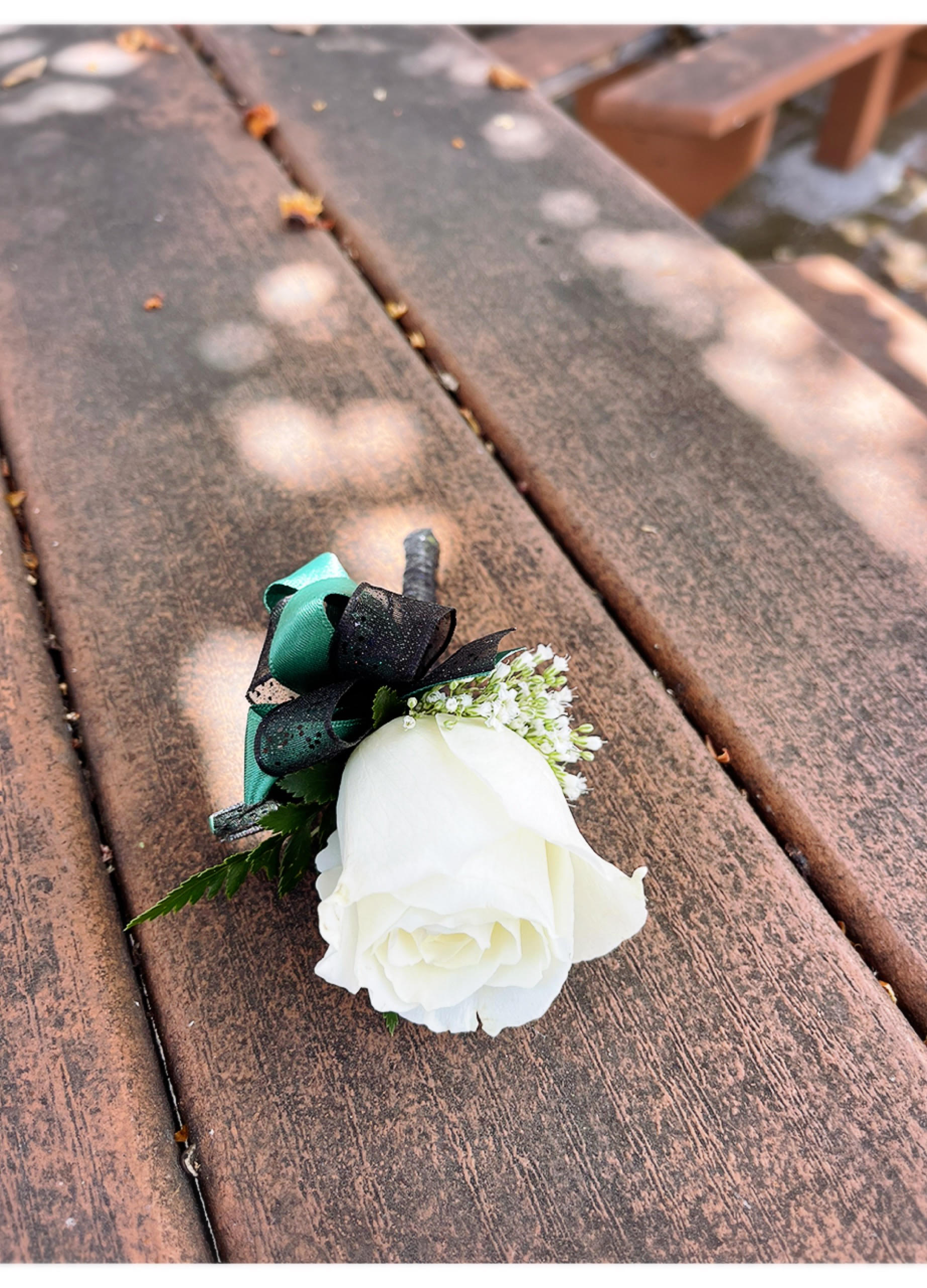 White rose boutonniere with black and teal ribbon on a wooden bench