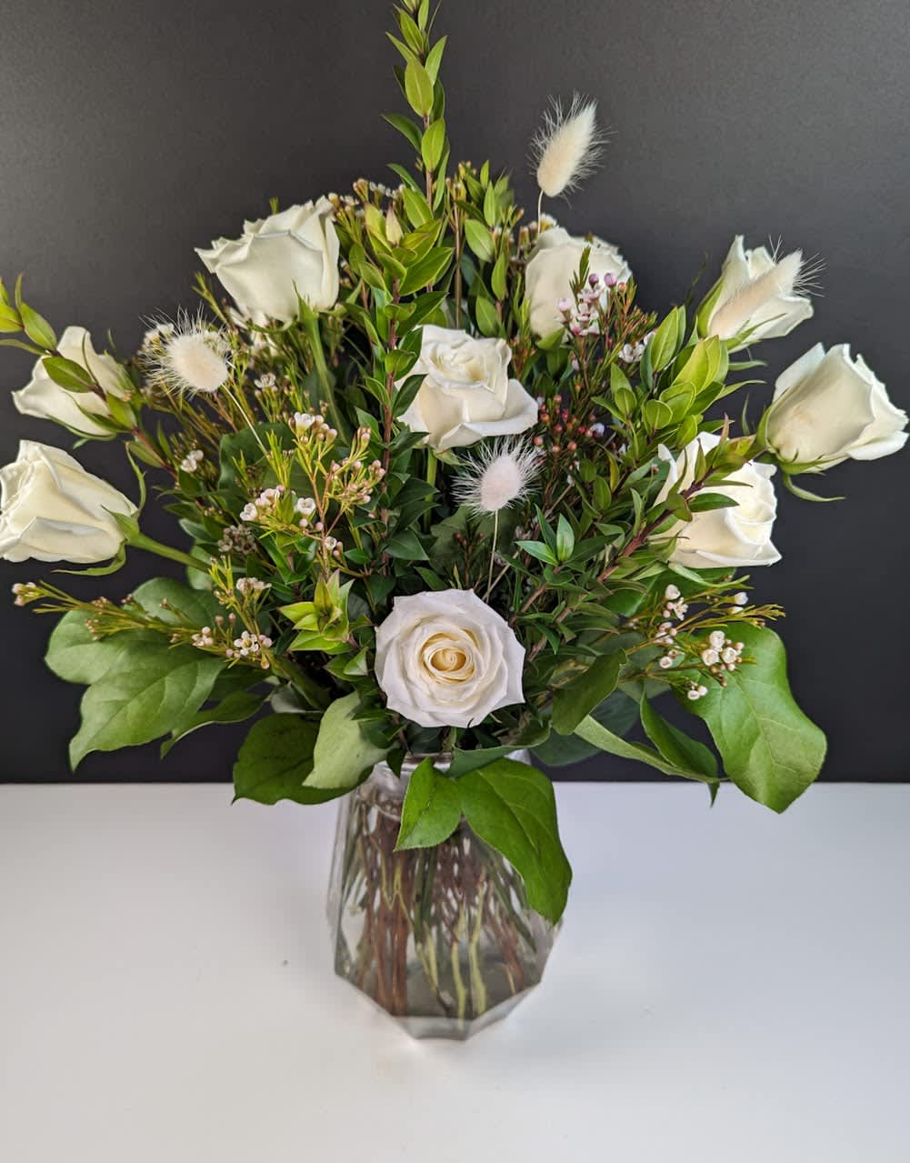 White roses arranged in a clear glass vase with greenery and small white accents