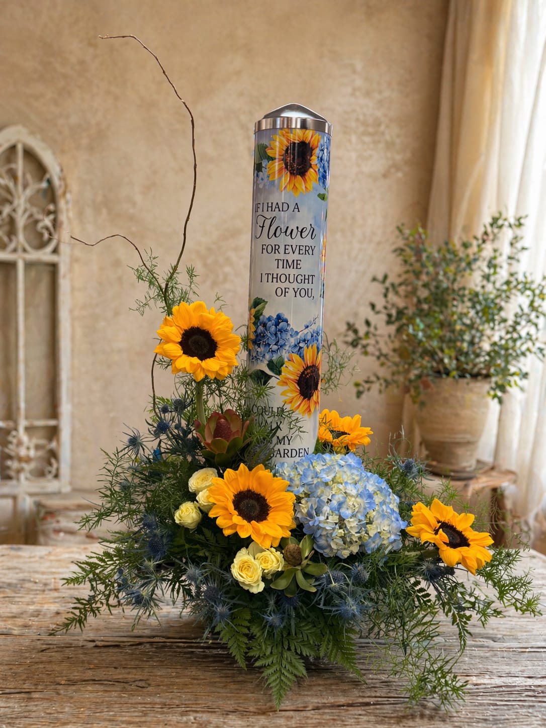Sunflower Rememberance  - A heartfelt tribute designed to honor cherished memories, Sunflower Remembrance blends vibrant sunflowers, soft blue hydrangea, delicate roses, and lush greenery in a natural, garden-inspired design. At its center stands a beautifully adorned keepsake art pole featuring a touching message, creating a lasting symbol of love and remembrance.  Accented with airy textures and graceful movement, this arrangement offers both comfort and beauty—perfect for memorials, sympathy gifts, or as a meaningful piece to brighten a loved one’s space. The combination of sunny blooms and calming blues evokes warmth, peace, and enduring connection. 