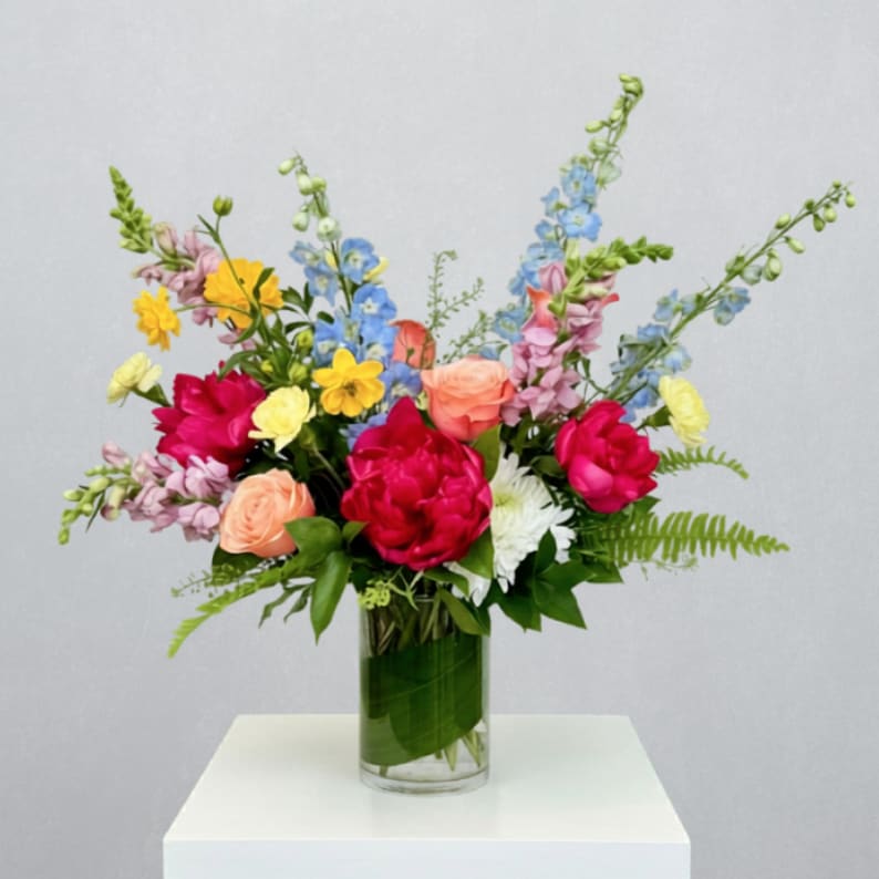 Mom's Favorite Flowers - MOTHER'S DAY COLLECTION | LOCAL DELIVERY ONLY  Celebrate Mom with a vibrant, joy-filled arrangement designed to feel as special as she is. Styled in a 5x8in cylinder with a fresh leaf wrap, this design features peonies, cremon mums, delphinium, snapdragons, short stem roses, butterfly ranunculus, carnations, and pennycress—creating a lush, colorful mix. With its bright, cheerful palette and garden-inspired movement, this arrangement is the kind of gift that will make Mom pause, smile, and feel truly appreciated.