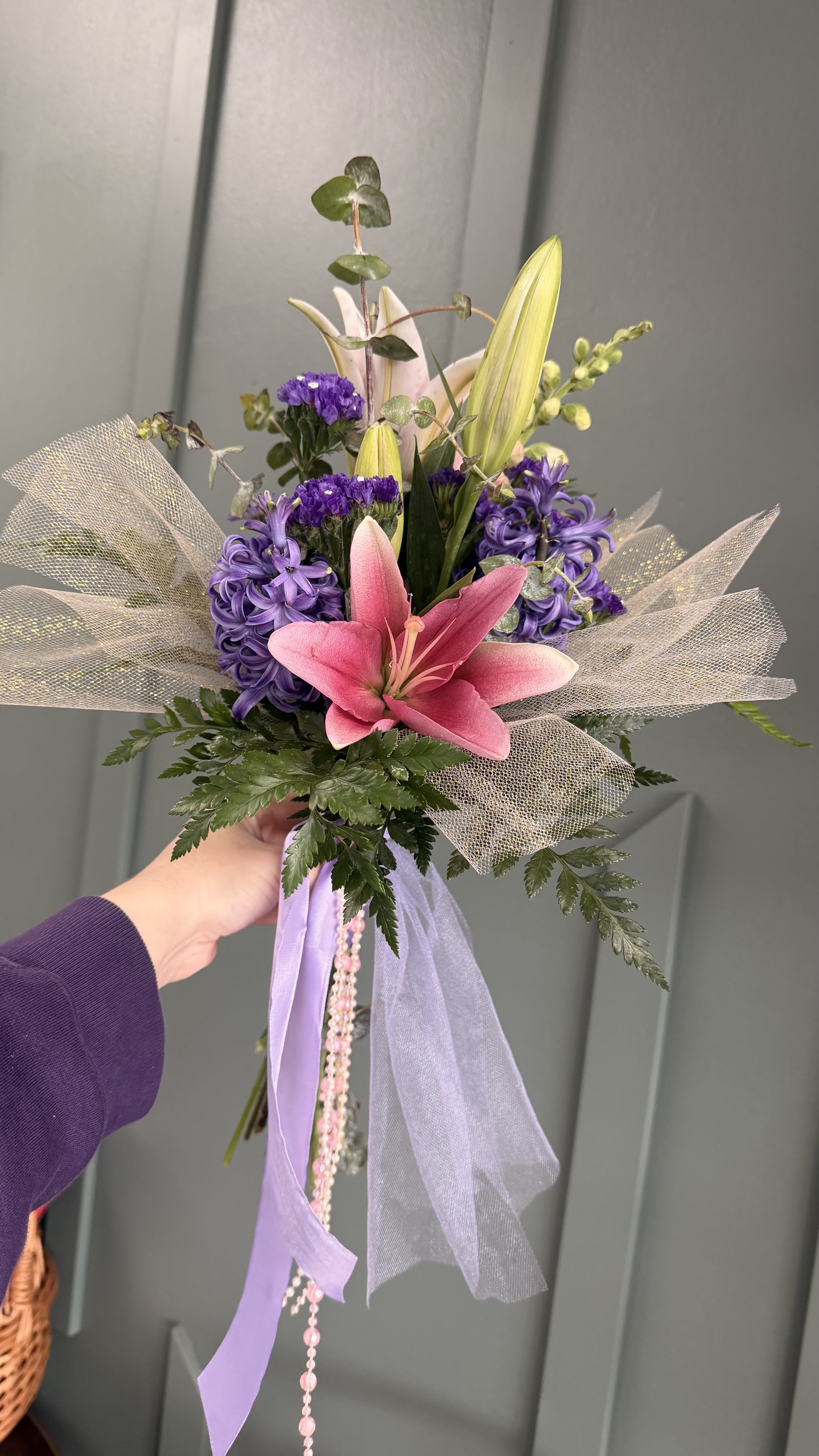 Prom Bouquet - A single large pink lily takes center stage in this prom bouquet, creating an instantly romantic focal point perfect for prom or other special events. The hand-tied arrangement reads youthful and whimsical-purple clustered blooms and pale lavender hyacinth accents surround the central lily while unopened lily buds add a fresh, hopeful touch-so the person you're giving it to will look and feel celebrated as they step into the evening.  Wrapped in airy tulle and finished with long lavender ribbons plus a delicate beaded strand, the bouquet's presentation is as important as the flowers themselves. Colors run from vivid pinks through soft lavenders and clean whites, giving the arrangement a lively, feminine palette that pairs beautifully with formalwear. Its compact, handheld size makes it ideal for carrying during a dance or posing for photos; light misting and keeping stems in water until the last moment will help the lilies and hyacinth stay fresh through the night.