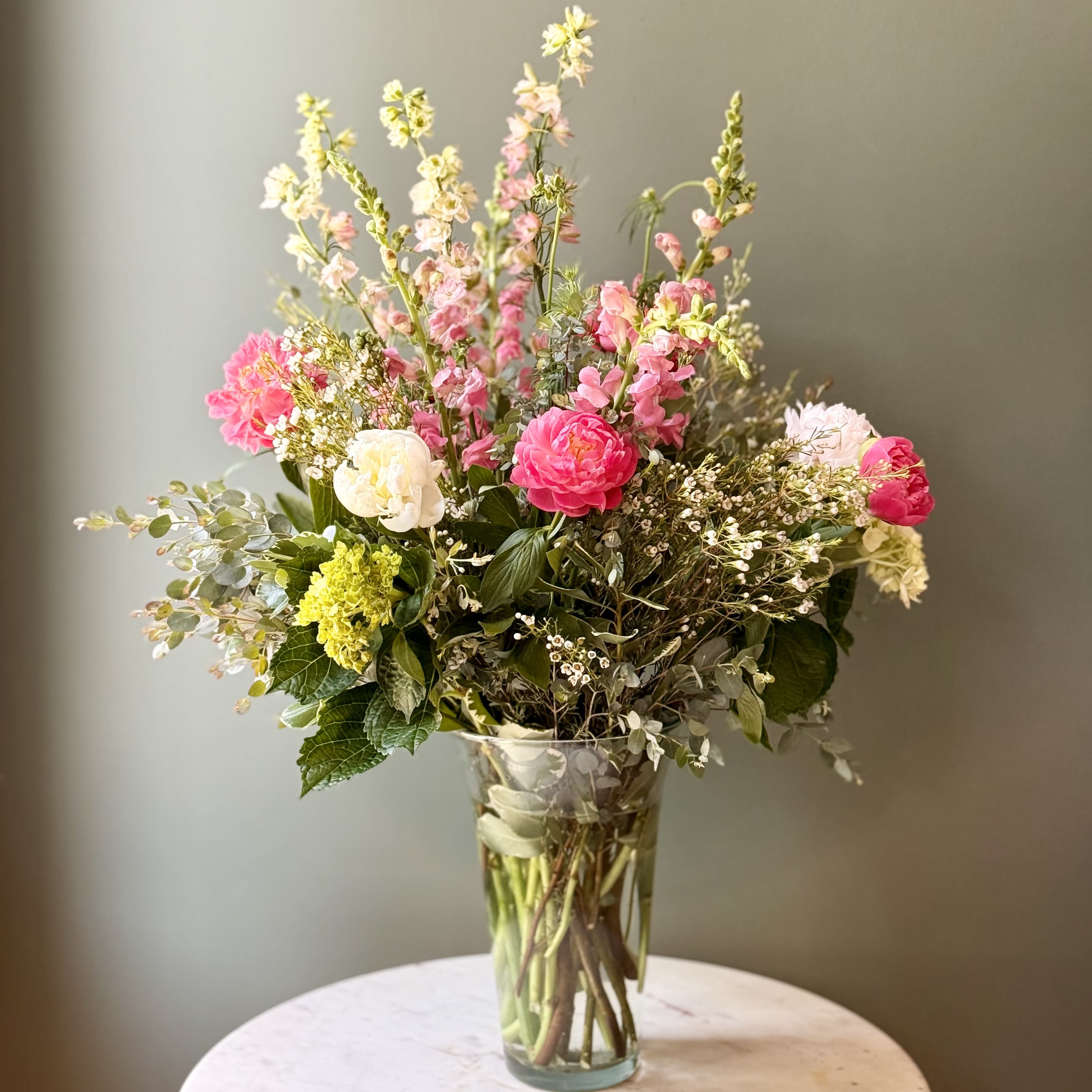 Garden-Inspired Peony - Garden-Inspired Peony Floral Design is a fresh, romantic peony arrangement crafted to capture the natural beauty of a blooming garden. Featuring: - Locally Grown Peony - Pink Snaps - Mini green Hydrangeas - Larkspur - Wax Flower - Styled with premium seasonal foliage Presentation Options: Tall clear vase measuring approximately 11". Vase included in price. Every order includes: A custom message card - simply enter your message at checkout and we'll take care of the rest. * pPlease note, Peony color may vary depending on availability.