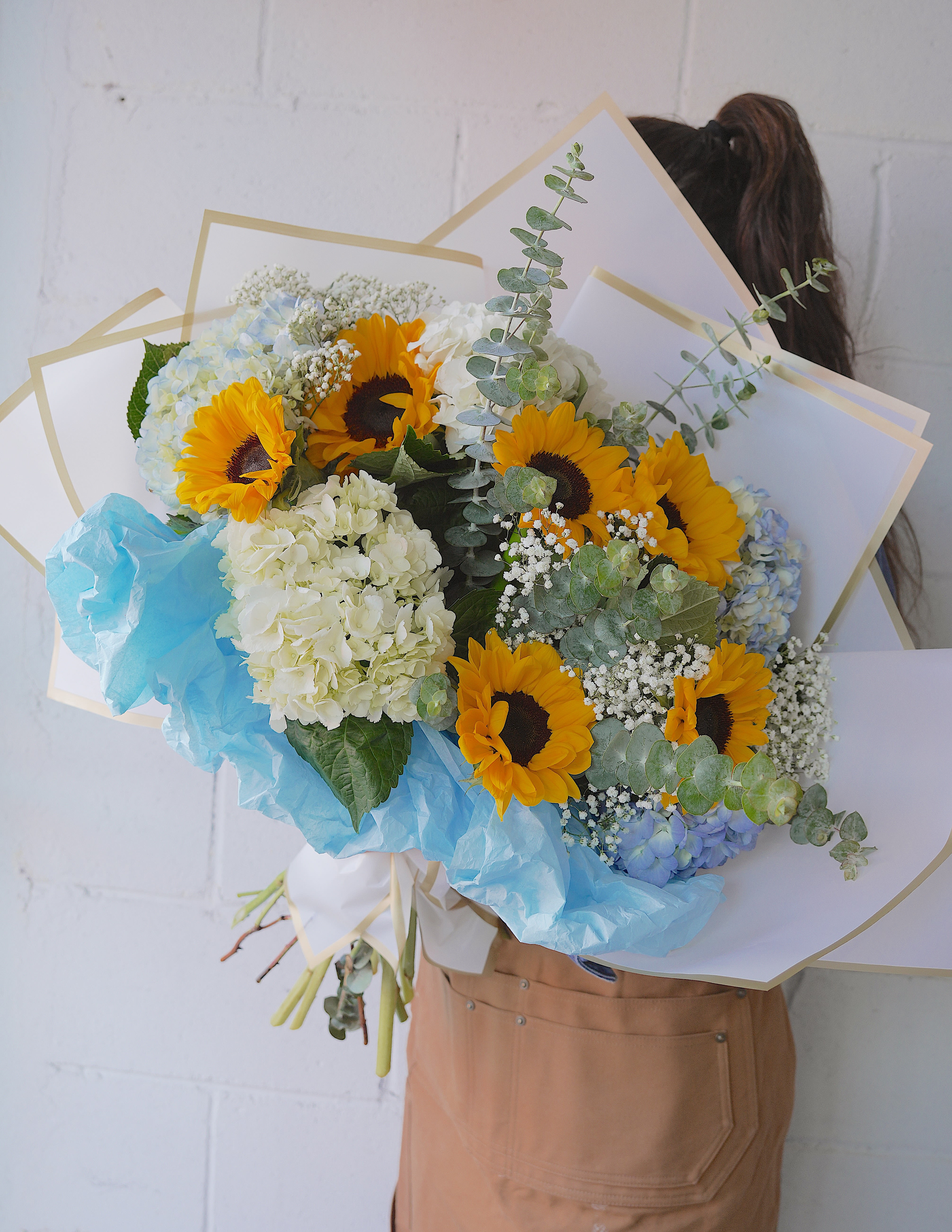 SUNFLOWER BREEZE - Bright sunflowers set a joyful tone in this sunflower and hydrangea bouquet-a sunny birthday bouquet or cheerful congratulations arrangement that instantly brightens the person you're thinking of. The combination of vivid yellow blooms with white and pale blue hydrangeas creates an uplifting, slightly romantic look that reads fresh and springlike, perfect for birthdays, thank-yous, or "just because" moments when you want someone to feel truly seen.