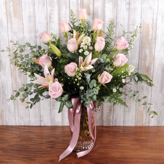 Let's Spoil Mom - Pale pink roses and open pink lilies take center stage in this Mother's Day and birthday flower arrangement, a gentle gift that will make the person you're thinking of feel truly cherished. The airy cluster of small white daisy-like blooms and eucalyptus gives the bouquet a soft, garden-style silhouette that reads romantic and fresh-perfect for anniversaries or a just-because "let's spoil Mom" moment.  Arranged in a clear glass vase and finished with a long pink satin ribbon, the display stands tall and open rather than dense, so the blossoms breathe and each stem shows off its texture and scent. Colors are a soothing mix of pinks, whites, and green foliage; lilies add drama and fragrance while roses bring classic warmth and the daisies add a cheerful, delicate touch.  This is the kind of bouquet that makes her feel seen-warm, celebrated, and loved-whether you're marking Mother's Day or surprising her on an ordinary Tuesday. Vase included; to keep flowers looking their best, refresh water daily and trim stems every few days.