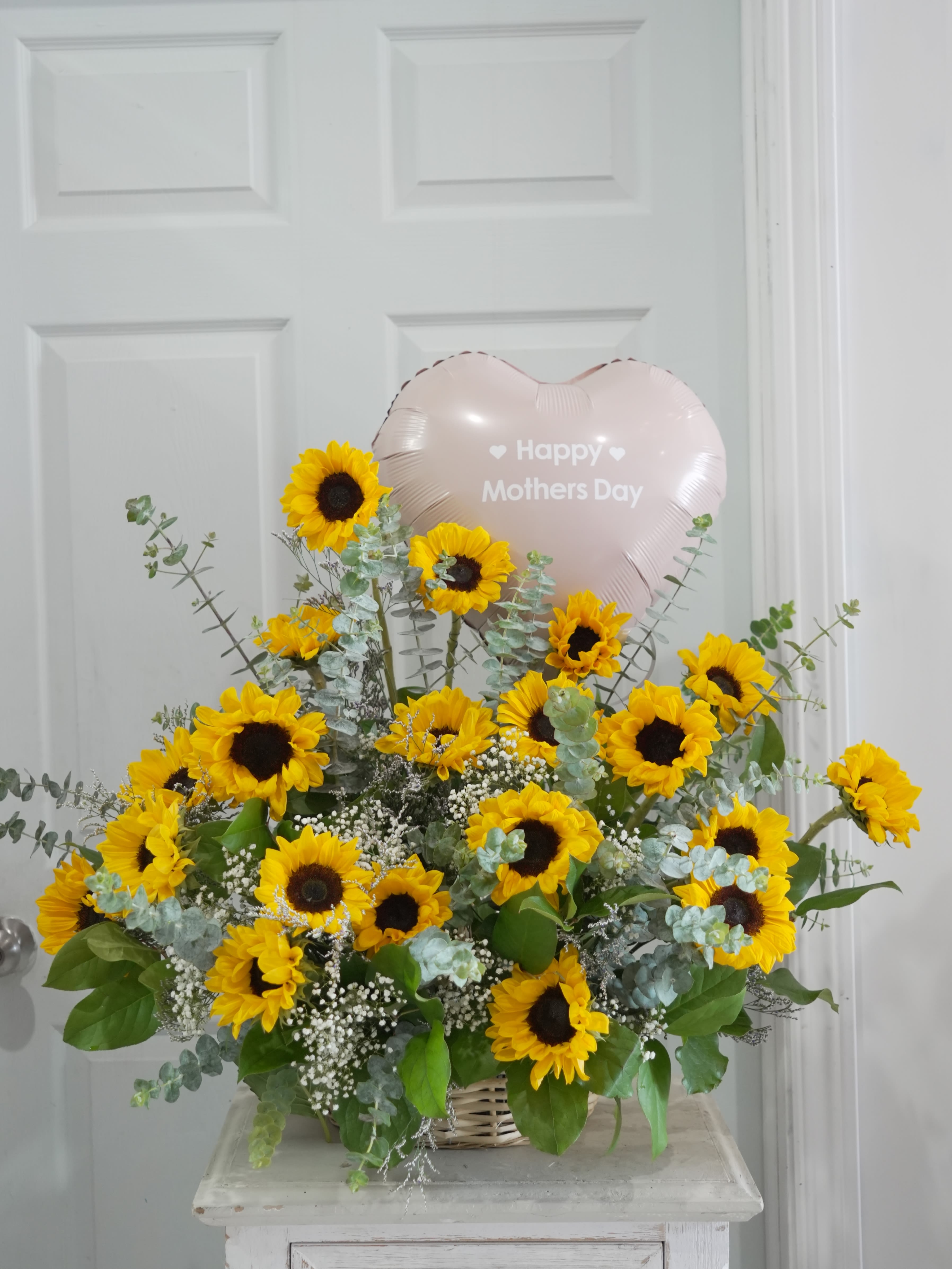 SUNFLOWER  GARDEN BASKET - Bright yellow sunflowers with white fillers and silvery-green eucalyptus in a woven basket, topped with a pink heart balloon-cheerful, heartfelt and perfect for Mother's Day or a sunny thank-you.