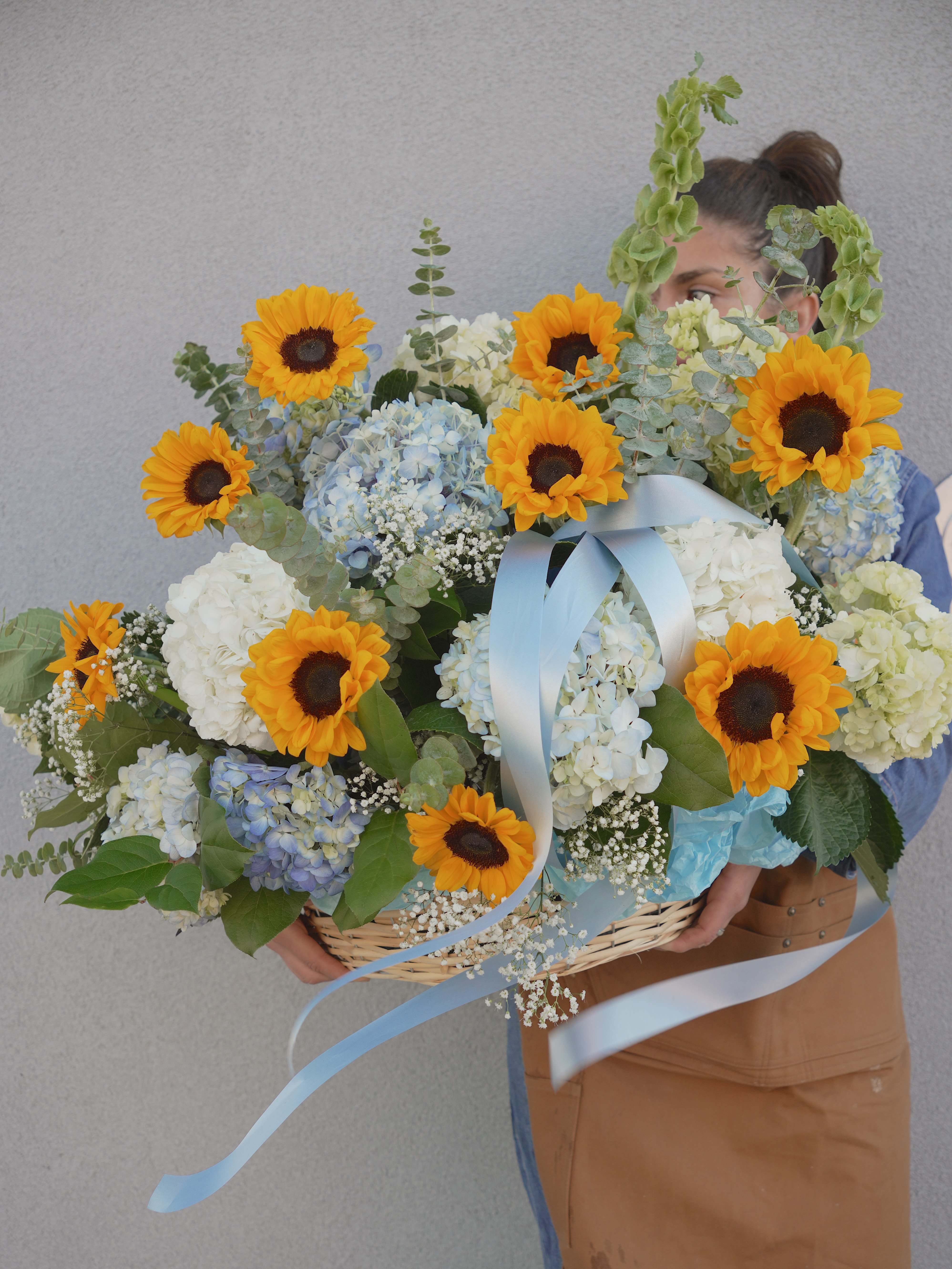 THE HAMPTONS BLOOM - Bright yellow sunflowers take center stage in this cheerful flower arrangement, paired with pale blue and white hydrangeas for a soft, fresh contrast-perfect as a birthday bouquet, congratulatory gift, or springtime surprise. The mix of bold sunflower faces and cloud-like hydrangea clusters reads as joyful and abundant, so the person you're sending it to will feel genuinely celebrated and uplifted.