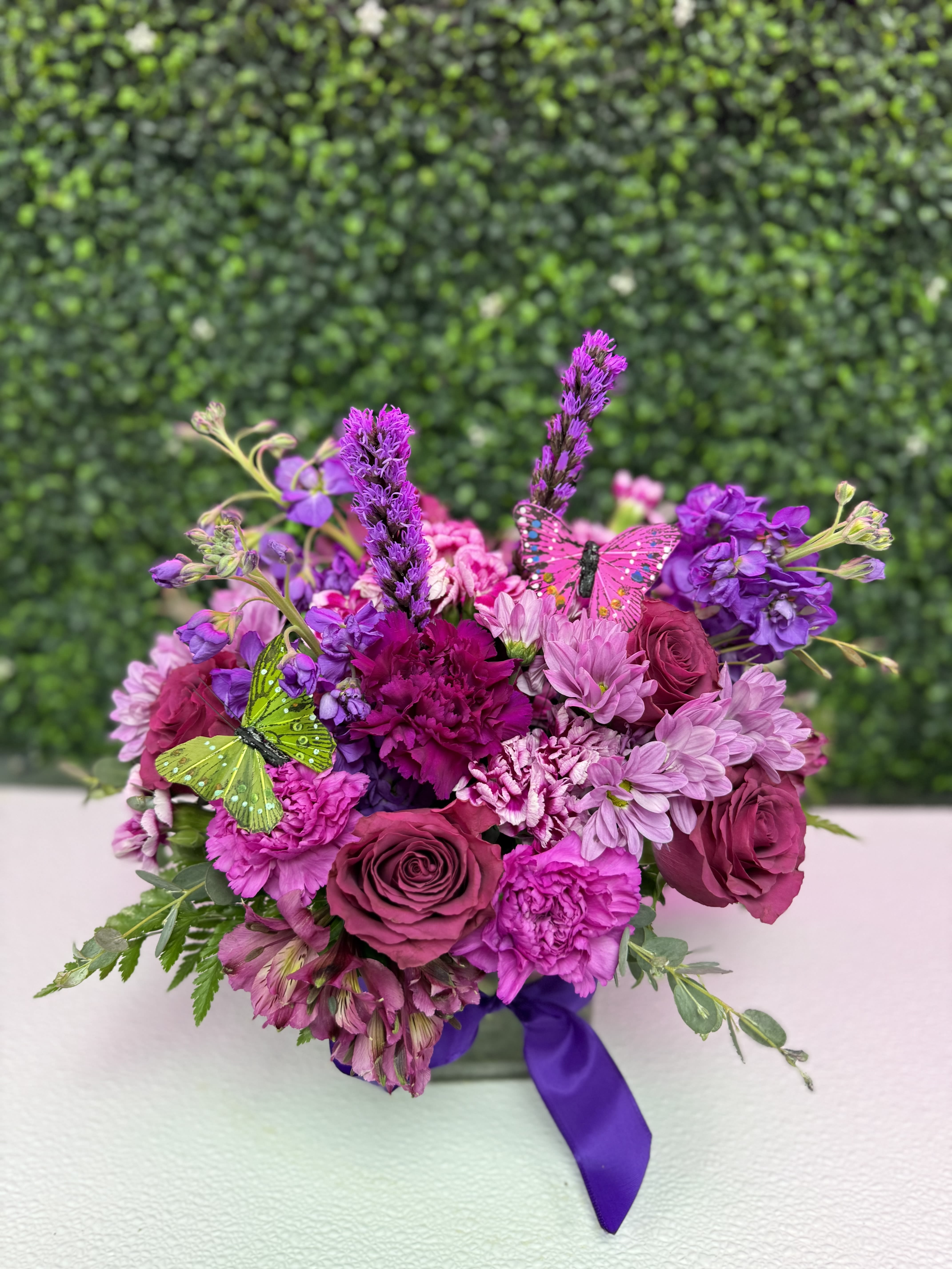 Mandy's Lavender Sky  - Lavender-leaning pinks and purples take center stage in this hand-tied flower arrangement perfect for a birthday bouquet or to say "I'm thinking of you." Mandy's Lavender Sky blends deep magenta roses and pink daisy-like blooms with purple carnations and tall lavender accent spikes to create a layered, textured look that reads both romantic and cheerful-the kind of bouquet that makes the person you're sending it to feel noticed and adored.  Soft white and green touches brighten the mix while decorative butterfly picks add a whimsical finishing note. The bouquet arrives finished with a purple ribbon, compact and easy to display on a bedside table or desk. Sized to give presence without overwhelming a small space, it's a fresh, fragrant choice for anniversaries, congratulations, or a meaningful pick-me-up.