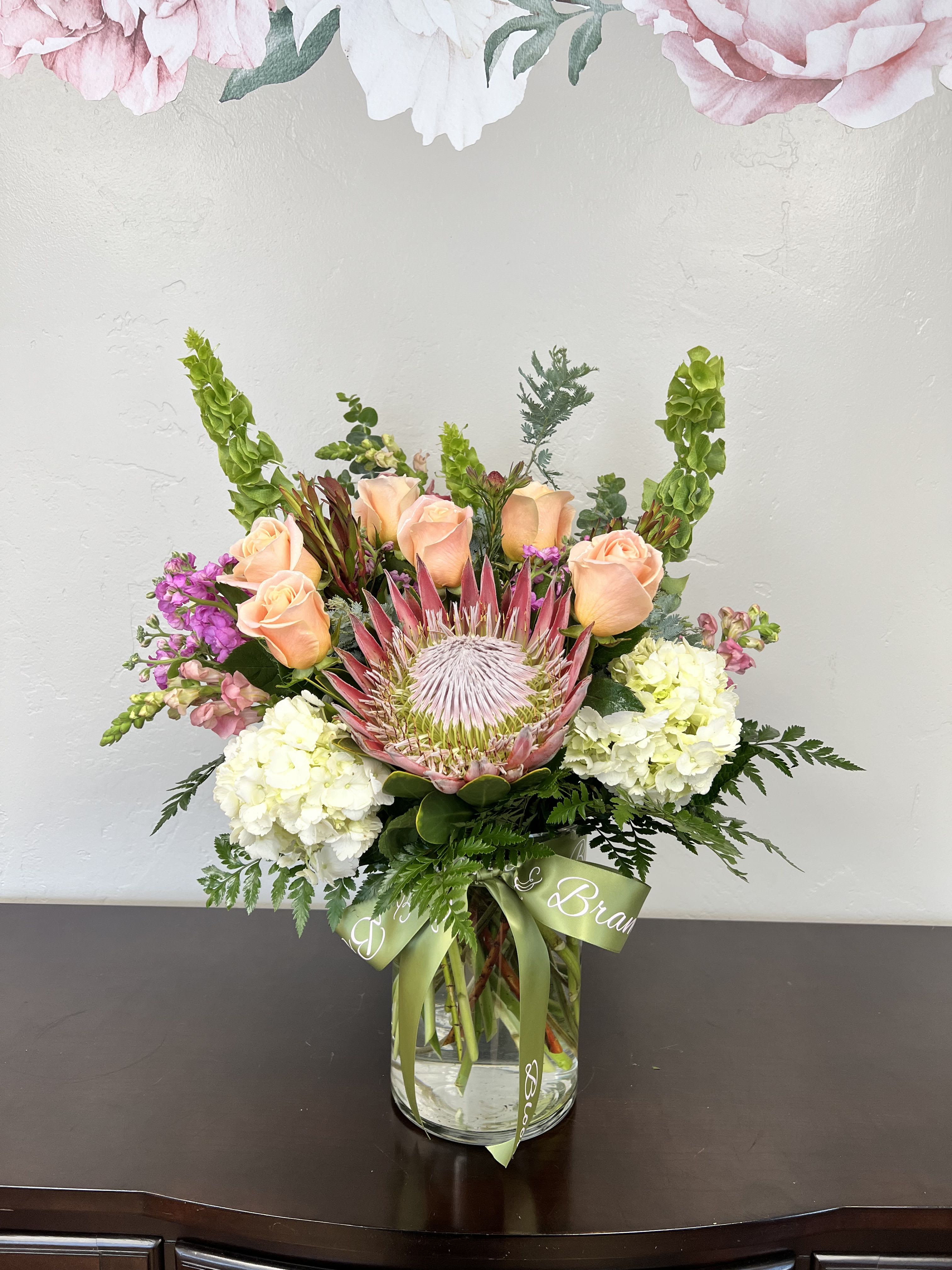 The Best Mom - The Best Mom! A bold pink protea crowns this bouquet, creating a striking focal point in a Mother's Day or birthday flower arrangement that feels both modern and heartfelt. This "Mom" arrangement reads as a polished everyday gift for the person you want to honor-perfect for birthdays, thank-you gestures, or spring celebrations-so she'll feel seen and celebrated the moment the vase is set down.  Surrounding the protea are soft peach roses, creamy white hydrangea clusters, and vertical pink snapdragon spikes, with tall green accents and airy foliage that give the bouquet a layered, garden-fresh silhouette. The clear glass vase and simple green ribbon keep the look clean and elegant while letting the pink, peach, white, and green palette shine. The composition is full and balanced, offering visual texture from the protea's dramatic form to the hydrangeas' cloud-like softness.  This arrangement arrives ready to display in the included vase-an effortless way to brighten a kitchen table or bedside-making it an ideal pick when you want to say "thank you," "happy birthday," or "thinking of you" with floral charm. Standard care: keep water fresh and trim stems every few days to extend the life of the roses and hydrangeas; the protea's sturdy bloom will maintain its presence as the arrangement evolves.