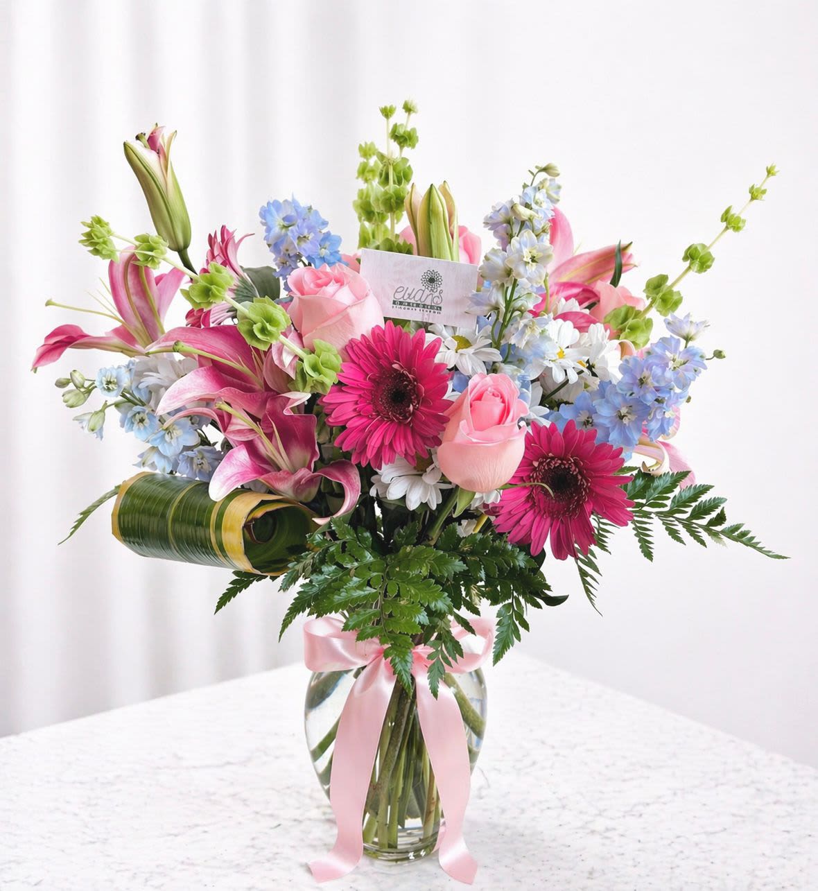 Mom's Meadow - This crystal vase arrangement has a mix of blooms, representing a mother's ability to nurture many things at once. This arrangement comes with Gerber daisies, pink roses, bells of Ireland, delphinium, and lilies. 