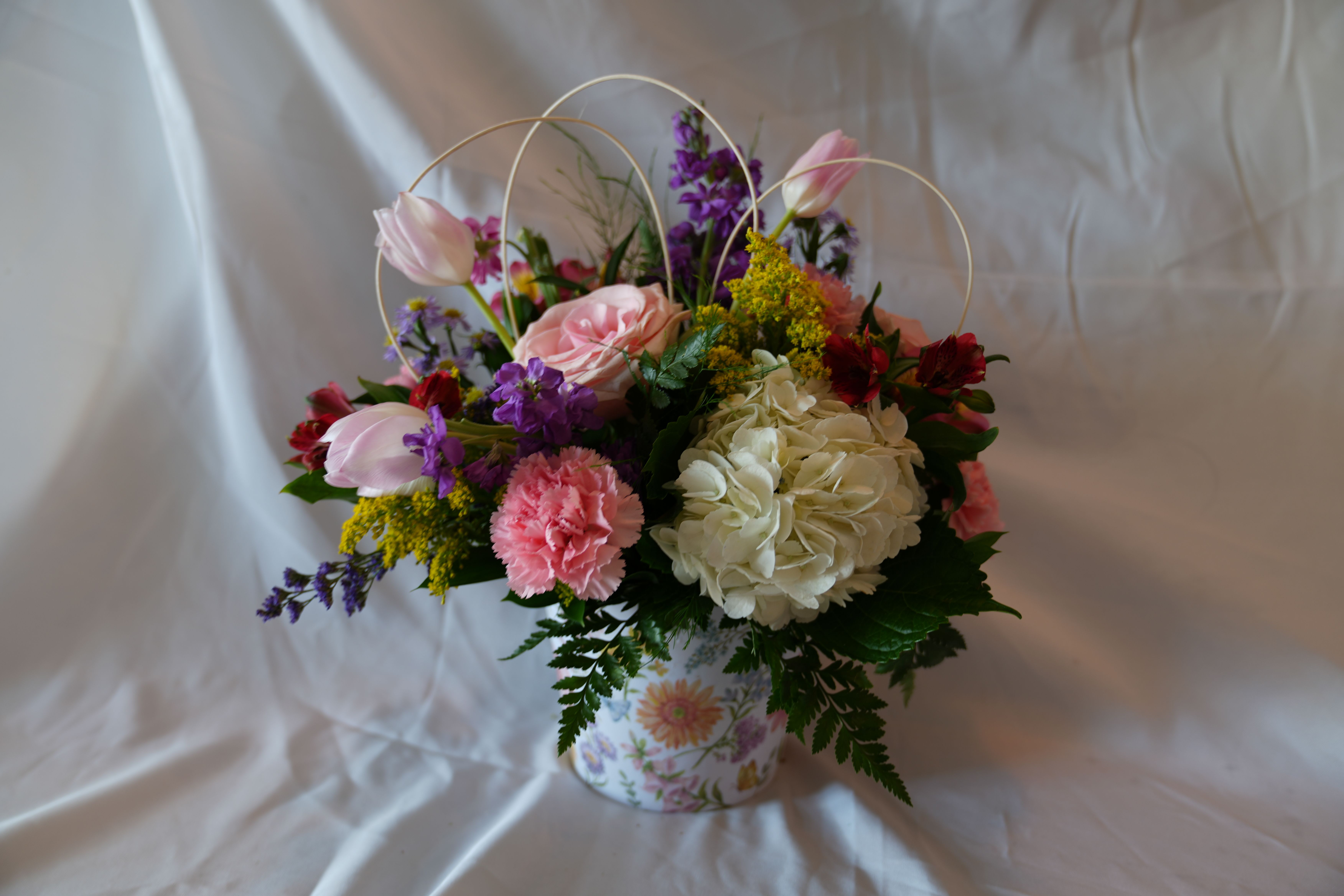 Garden of Gratitude - A cheerful mix of pink, white, purple, and yellow blooms makes a sweet statement for birthdays, Mother's Day, or to say "thank you" with warmth - the kind of flower arrangement that will make the person you're thinking of feel genuinely seen. Garden of Gratitude is a floral arrangement designed as a giftable bouquet in a decorative floral-print tin, its arched wire handles giving a charming basket-like silhouette that reads as both playful and polished.