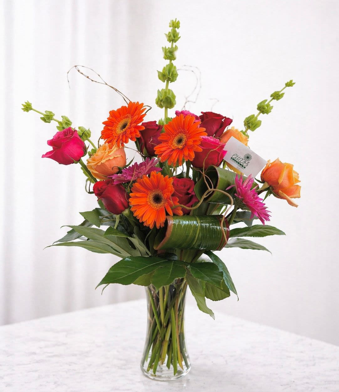 Mom's Radiance - This vase arrangement contains sun-like orange and pink gerberas, bells of Ireland, red roses, hot pink roses, orange roses, and tropical leaves. This arrangement is for the mom who has an bright and energetic presence.  